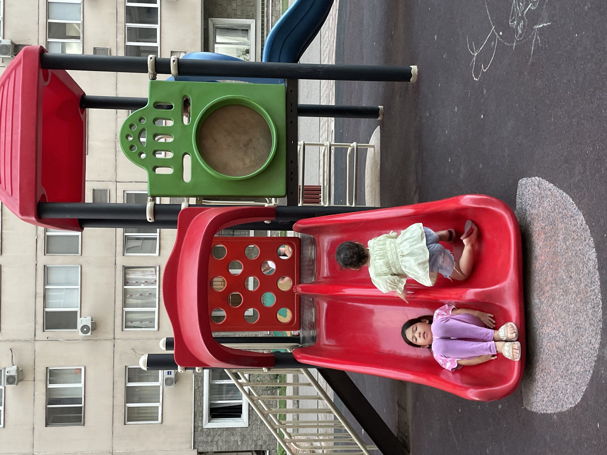 In Almaty, Kazakhstan, a red double-slide playground structure is the central focus. A young girl, wearing purple leggings and a white and pink striped shirt, sits at the bottom of the left slide, facing forward. To her right and slightly up the slide, another young girl, wearing a light green ruffled shirt and denim shorts, is positioned with her back to the viewer, appearing to be climbing or preparing to slide. The playground is situated on dark, speckled ground, with a chalk drawing of a face visible to the right. Behind the playground, a beige apartment building with numerous windows and air conditioning units stands. The scene is captured in daylight.