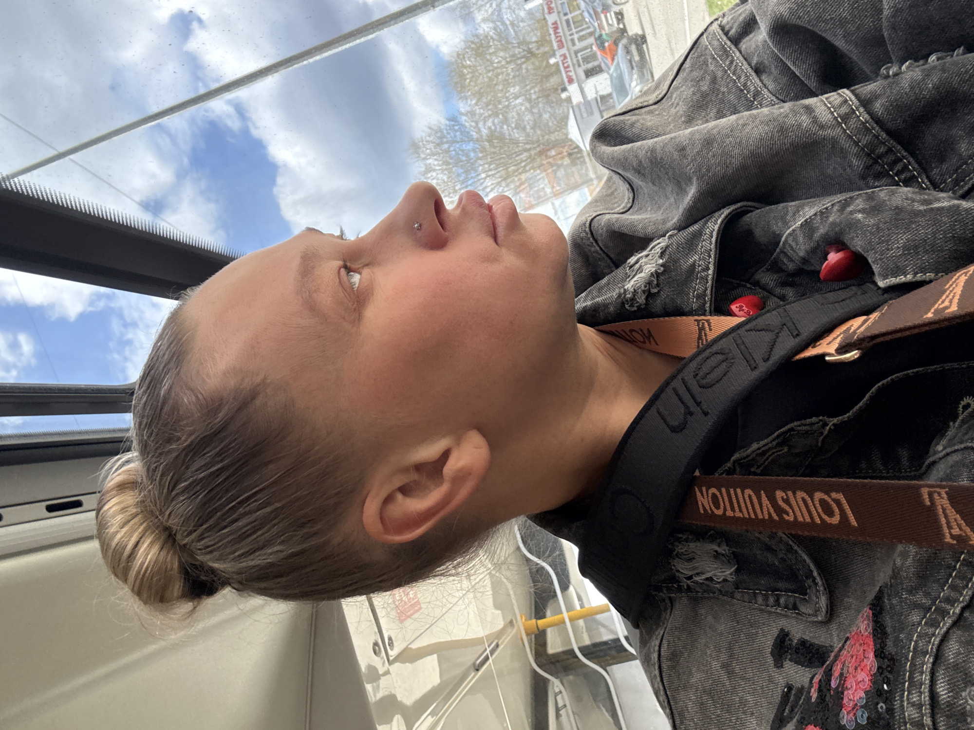 A side-profile image captures a person, likely female, with blonde hair tied in a neat bun, looking upwards and out of a window. A small silver stud piercing is visible on their nose. The person is wearing a dark, distressed denim jacket that features some red sequined or embroidered details. Tucked into the jacket's folds are two small, bright red heart-shaped objects, possibly earplugs or decorative items.
Across the person's shoulder, a black strap with the text "Calvin Klein" (partially visible) and a brown strap with the inverted text "LOUIS VUITTON" are visible, suggesting branded accessories or a bag.
The setting appears to be inside a vehicle, such as a bus or tram, looking out a large window. The outside view shows a partly cloudy sky and reflections of urban elements, including buildings and bare trees. Some Cyrillic text, partially legible as "УРУТЫ ЩАРЫНА" or similar, is reflected in the window, indicating a Russian location. Given the provided context, this scene is set in Perm, Russia. The image was taken during daytime, characterized by clear natural light. The person's expression is neutral, seemingly focused on the view outside.