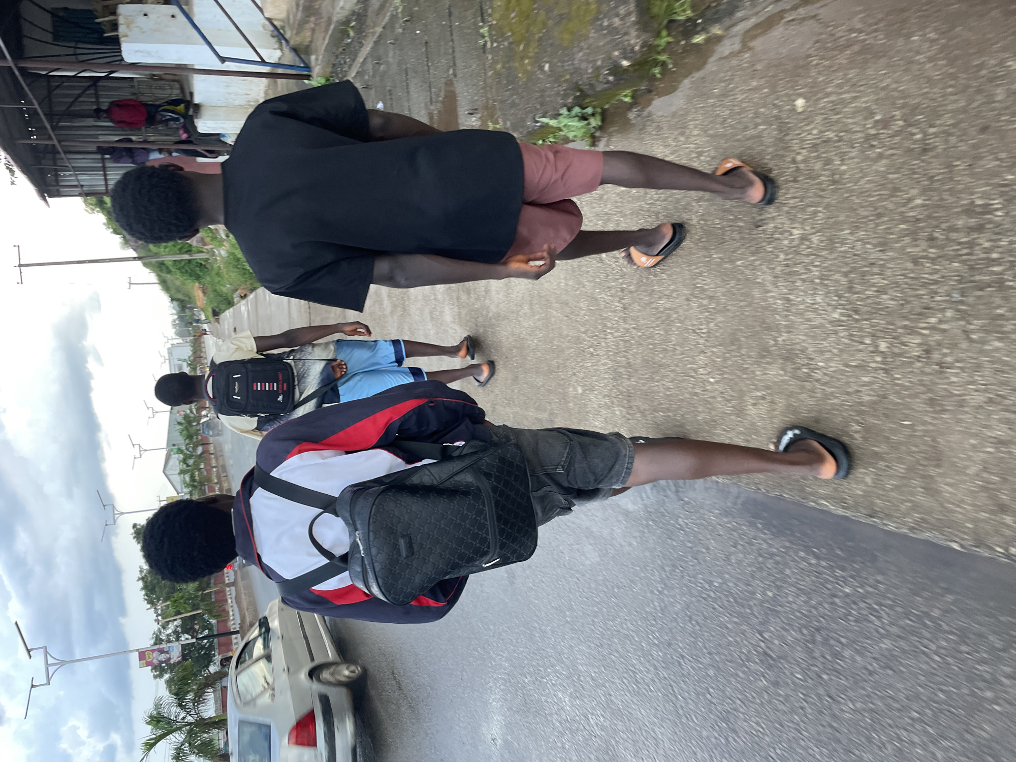 Three young males walk south along a paved sidewalk next to a busy street. The male in the foreground on the left wears a blue, white, and red jacket, denim shorts, and a black backpack. He walks on the sidewalk, facing away from the camera. To his right, the middle male wears a light-colored shirt, blue shorts, and a black backpack with red trim. He walks on the sidewalk, facing away from the camera. On the far right, the third male wears a black t-shirt, reddish-brown shorts, and orange slide sandals. He walks on the sidewalk, facing away from the camera. The street to the left is wet, suggesting recent rain. A silver sedan is visible on the street. Streetlights with solar panels are mounted along the street. Buildings are visible in the background on the right side of the image. The scene appears to be an urban environment in Jos, Nigeria.