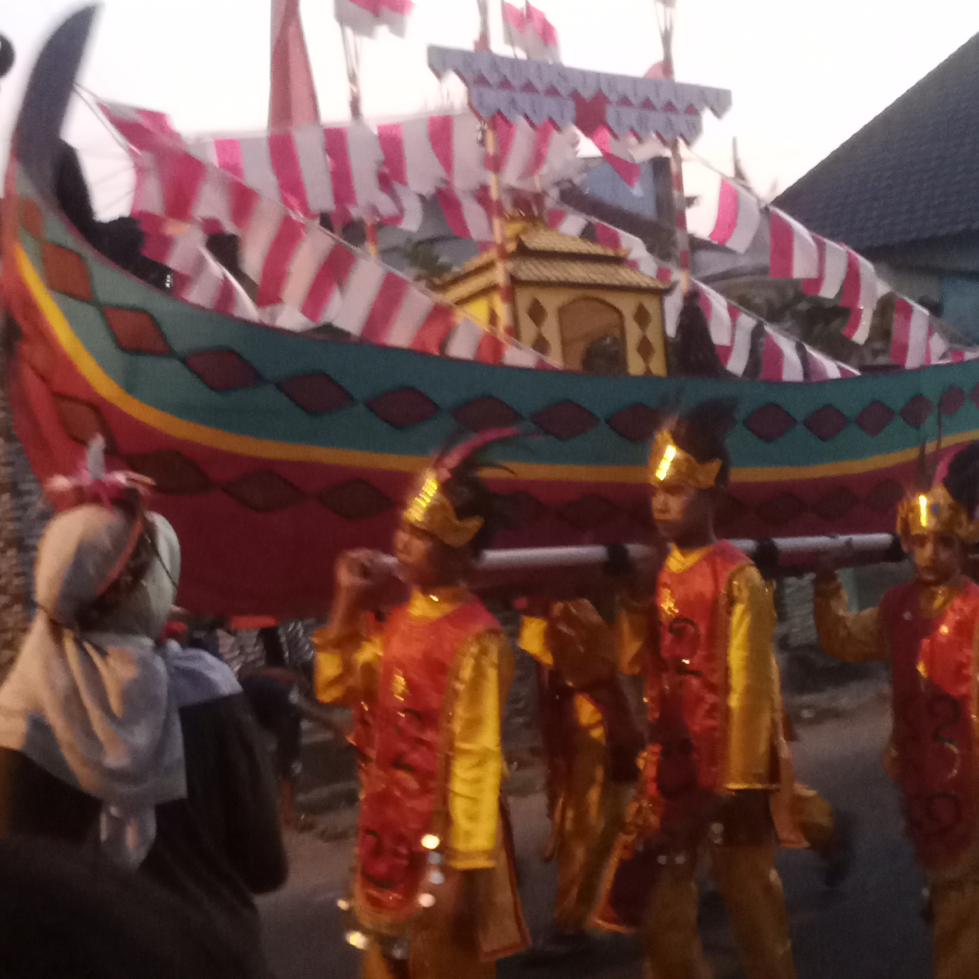 In Semanding, Indonesia, a group of people are participating in a procession. A large, decorative boat-like structure, adorned with red and white flags, is being carried. Several individuals, appearing to be young males, are dressed in traditional, ornate red and gold costumes and golden headwear, holding poles that support the boat. To the left, a person is wearing a grey headscarf and dark clothing. The procession is taking place outdoors, with a blurred background suggesting residential buildings and a darkening sky, indicative of late afternoon or early evening. The overall scene depicts a cultural event or parade.