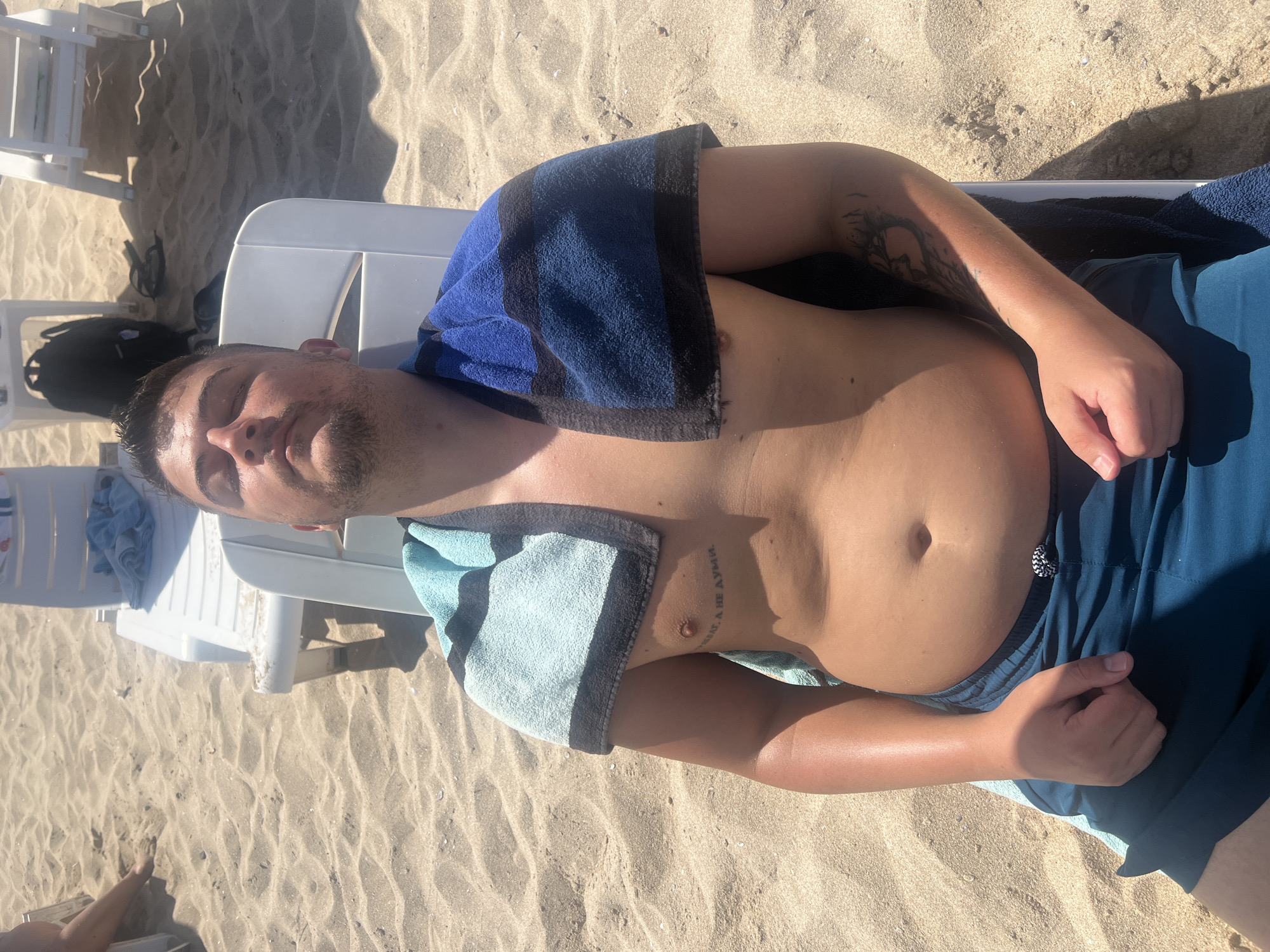 A young man with dark hair and a beard is lying on a white lounge chair on a sandy beach. He is wearing blue swim trunks and has his eyes closed, with a light blue and black striped towel draped over his shoulders and a dark blue and black striped towel on his lap. A tattoo is visible on his chest and another on his left arm. The sand is light brown, and in the background, there are other lounge chairs and some shadows cast by the sun. The image appears to be taken in Primorsko, Bulgaria, a popular seaside resort town.