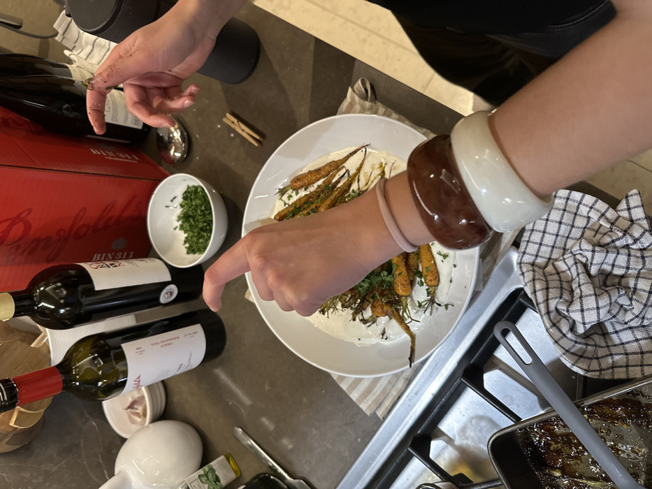 A high-angle shot depicts a pair of hands preparing food. One hand gestures toward a white plate containing a creamy base topped with roasted carrots and herbs. Nearby, a small bowl holds chopped herbs, and a case of red wine sits alongside several bottles. The scene unfolds on a countertop next to a stovetop. The person wears multiple bracelets on their wrist.