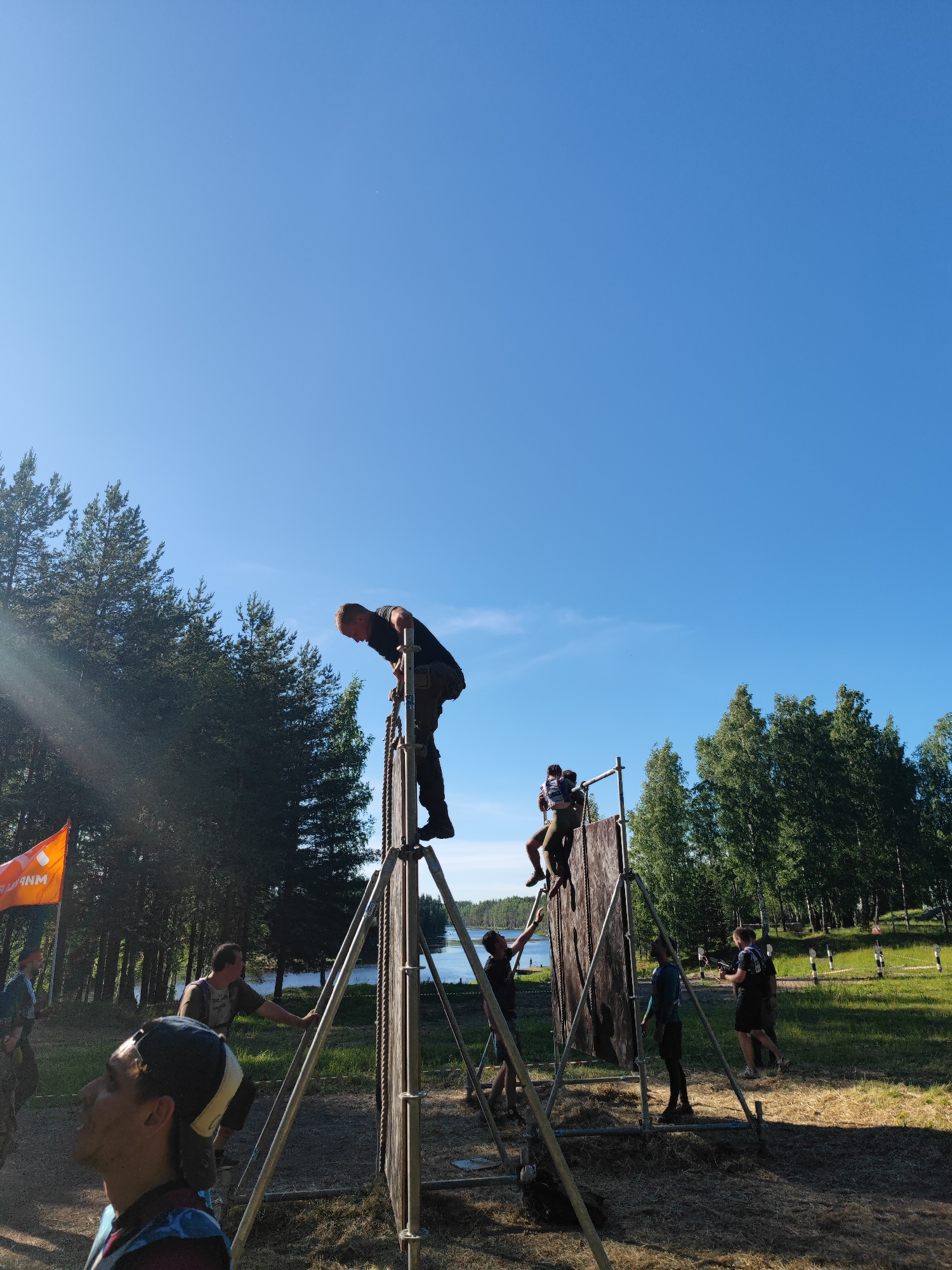 This is a low-angle, medium shot photograph taken outdoors on a sunny day, featuring a group of people participating in an obstacle course event. The background consists of a clear blue sky with scattered clouds and a line of green trees, indicative of a natural or park-like setting. In the middle ground, a large wooden wall obstacle is set up on a metal frame. Several individuals are interacting with this obstacle. One man, wearing a black t-shirt and dark pants, is positioned at the top of the wall, appearing to be in the process of descending. Another individual, in a lighter colored top and pants, is helping him from a slightly lower position on the obstacle. To the right, another person is climbing the wall, assisted by someone below. Further to the right, two other participants are visible, one in a black t-shirt and shorts, holding a rifle-like object, and another standing near him. In the foreground, a person wearing a baseball cap and a blue and black patterned shirt is looking upwards towards the obstacle course. An orange flag with white lettering is visible on the left side of the image, and a body of water can be seen beyond the obstacle in the background. The overall scene suggests a competitive or recreational outdoor event.