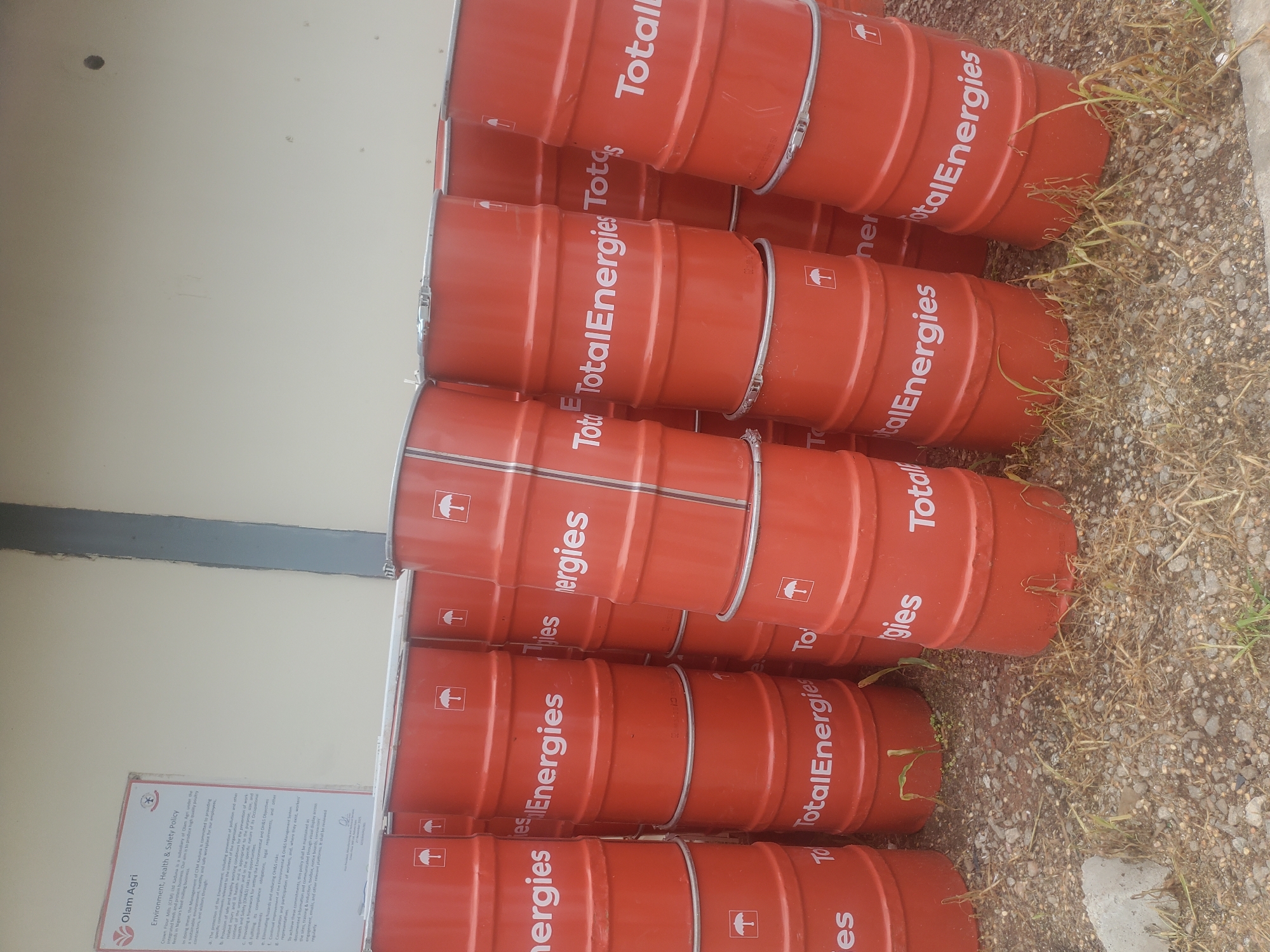 In the city of Gwagwada, Nigeria, a stack of bright orange, cylindrical barrels with white "TotalEnergies" branding and umbrella logos are arranged in uneven piles against a light-colored wall. The barrels are stacked several high, with some leaning precariously. Dry grass and gravel cover the ground around the barrels. A portion of a document with "Olam Agri" and "Environment, Health & Safety Policy" visible is also present in the upper left corner of the image.