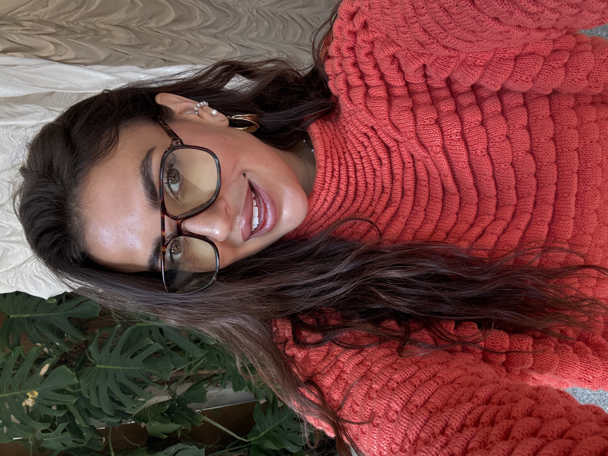 A woman with dark hair and glasses is pictured indoors. She wears a reddish-orange sweater with a textured pattern. In the background, there's a light-colored textured surface, possibly a curtain or bedspread. Green leaves, likely from a plant, are visible in the lower left corner. The scene gives no direct indication of being in Kuta Selatan, Indonesia; this location is not discernible from the image.