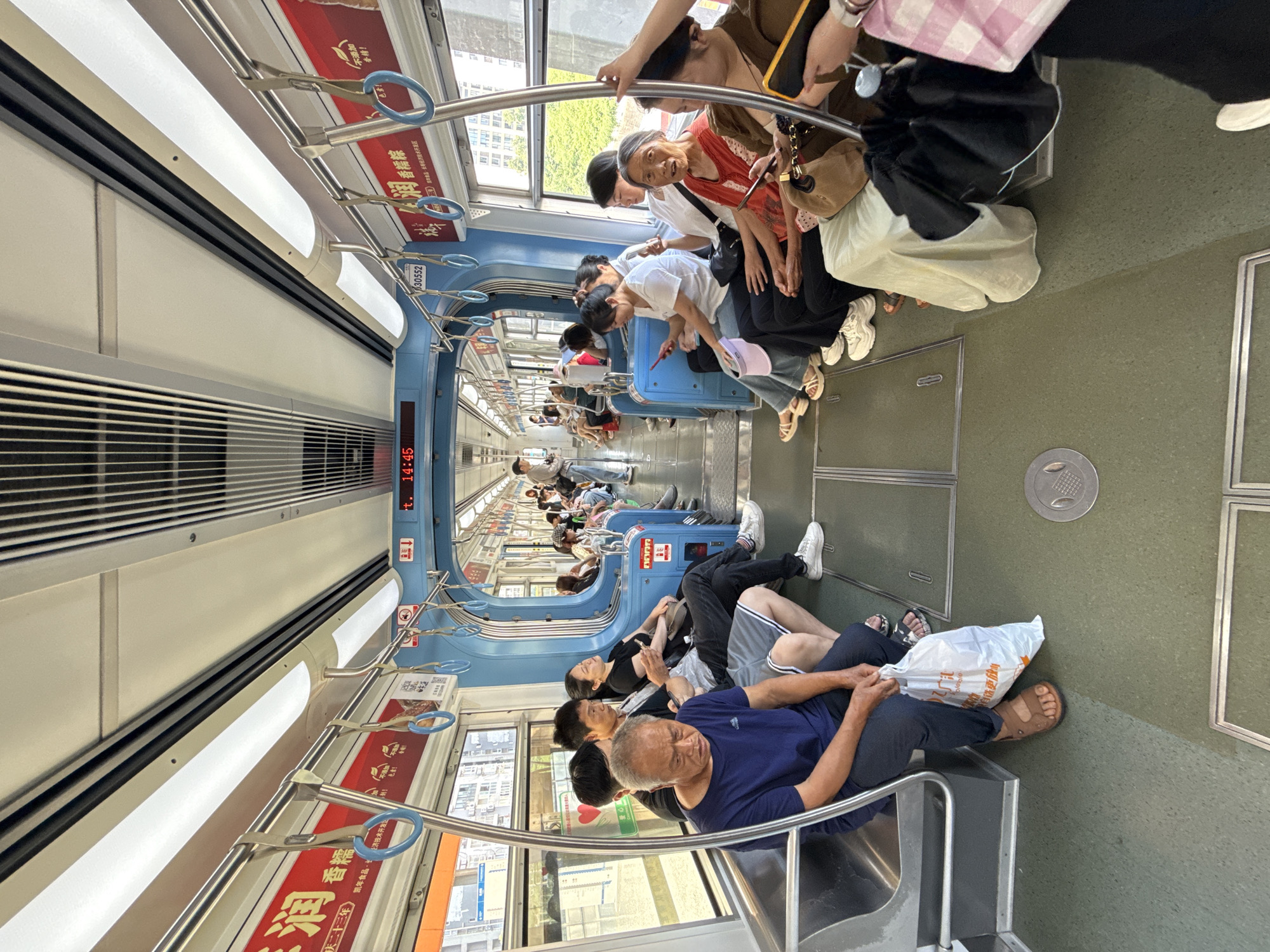 The image captures the interior of a subway car in Banan, China, heavily rotated clockwise. Numerous passengers are visible, either seated along the blue benches that line the windows or standing and holding onto blue-ringed handrails. The car appears moderately crowded.
In the foreground, an older man with grey hair, wearing a dark blue t-shirt and black pants, is seated, holding a white plastic bag. Next to him, other passengers are seated, some looking at their phones, others resting or conversing. Towards the mid-ground and background, more passengers are standing or seated further down the aisle, suggesting a typical commuting scene. An older woman in an orange top is looking towards the camera.
Notable details include the bright overhead fluorescent lighting, large windows revealing glimpses of buildings and greenery outside, and a green floor. A digital display screen is visible on the wall, showing the time as "14:45". Advertisements are prominently displayed above the windows.
Visible text on the advertisements includes: "润香格" (Rùn Xiāng Gé), "凯年食品、卡经济技术开发区" (Kai Nian Food, Card Economic and Technological Development Zone), "新!!" (New!!), and "不添加 香精!" (No added flavor/essence!). Another piece of text "30552" is seen on a panel. The overall mood appears calm, with people engaged in their journeys.