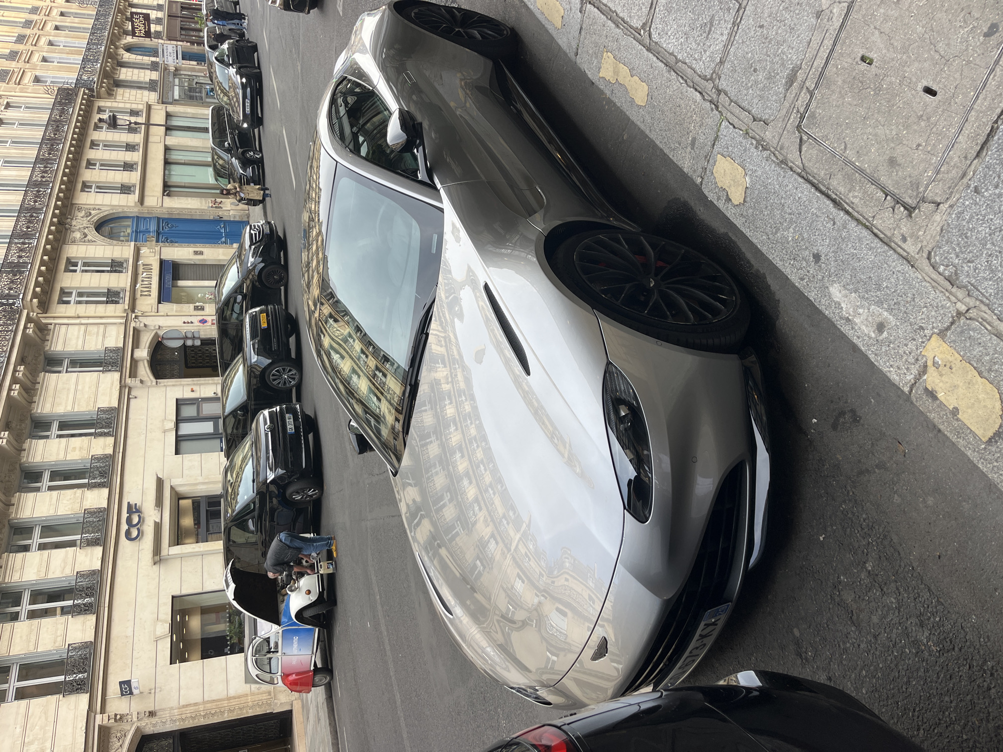 The image shows a silver Aston Martin parked on a street in Paris, France. The buildings along the street are typical Parisian architecture with ornate facades. Several black cars are parked behind the Aston Martin. A man is working on the engine of a red, white, and blue vintage Citroen 2CV. The building behind the Citroen has a "CCF" sign. The license plate of the Aston Martin visible to the left has the inscription "9673-KX". The street is paved with cobblestones.