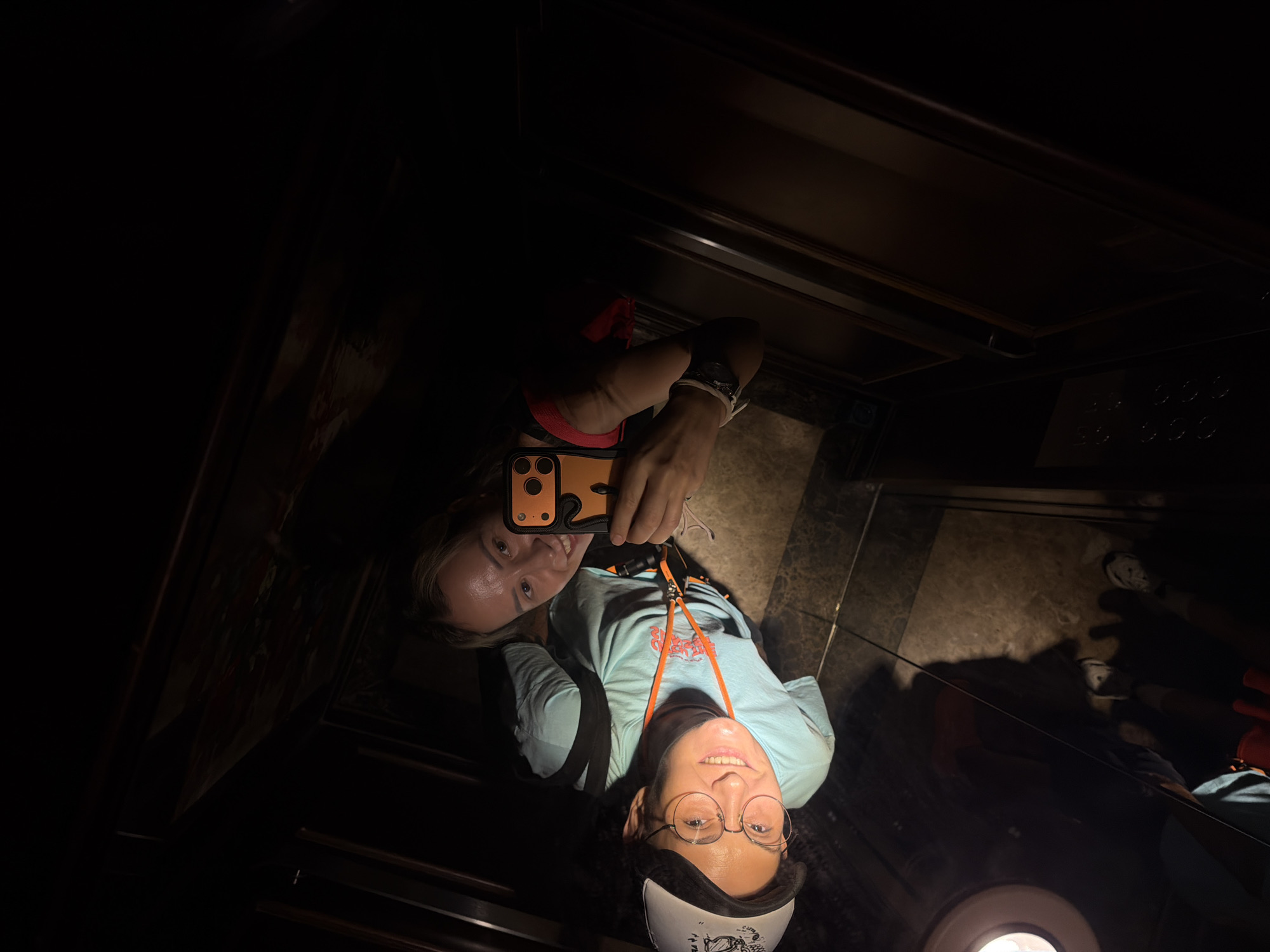 In a dimly lit elevator with mirrored walls, a woman is taking a selfie. She is wearing a light blue t-shirt, glasses, and a white hat. The elevator's interior features dark wood paneling and what appears to be a patterned carpet on the floor. The lighting is focused on the woman and her reflection, creating a dramatic effect. The surrounding environment is mostly dark, obscuring details of the elevator's exterior or location. There is no indication that this image is related to Thuận Hóa Ward, Vietnam.