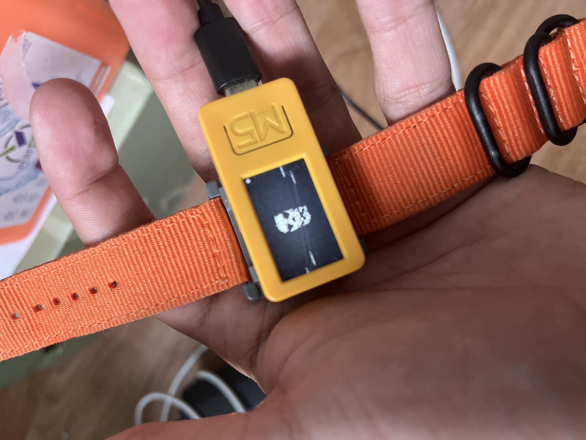A person's hand is holding an orange fabric watchband with a yellow electronic device attached. The device has a small screen displaying a pixelated image that resembles a video game. The watchband has several holes for adjustment and is secured with two black loops. A black USB cable is plugged into the bottom of the yellow device. The background is blurred, showing a wooden surface and a white electrical cord. There is no visible indication of the location being Belyye Berega, Russia.