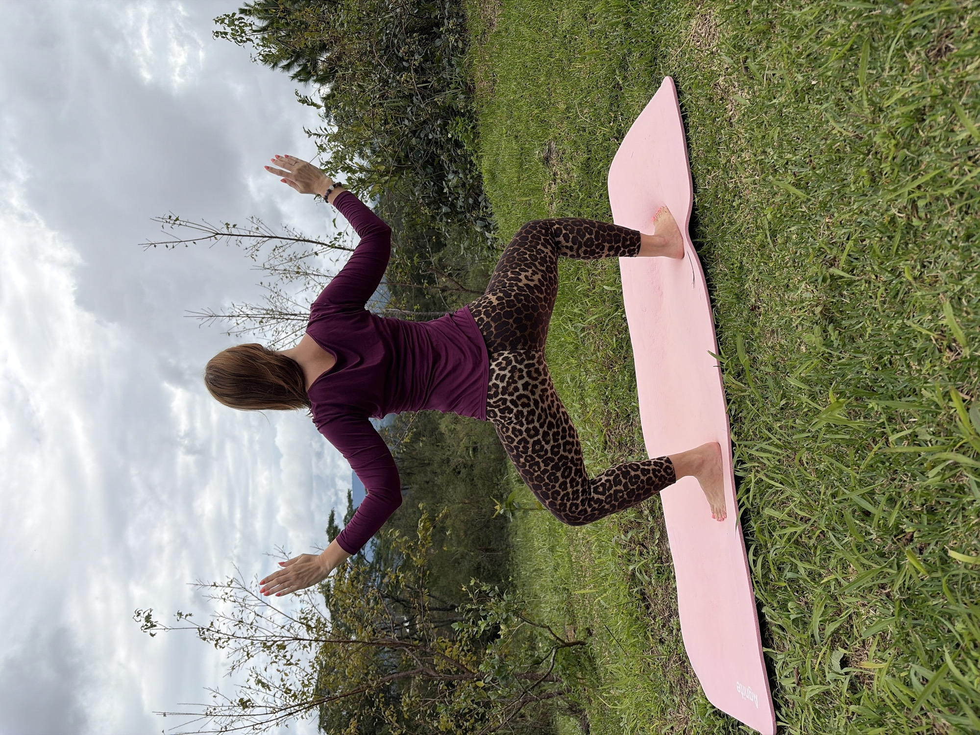 In Ban Khun Wang, Thailand, a woman with brown hair performs a yoga pose on a pink mat laid out on a grassy slope. She is facing away from the camera, wearing a plum-colored long-sleeved shirt and leopard-print leggings. Her arms are extended outwards, with her palms facing upwards. The background consists of trees and a cloudy sky, suggesting an outdoor setting.