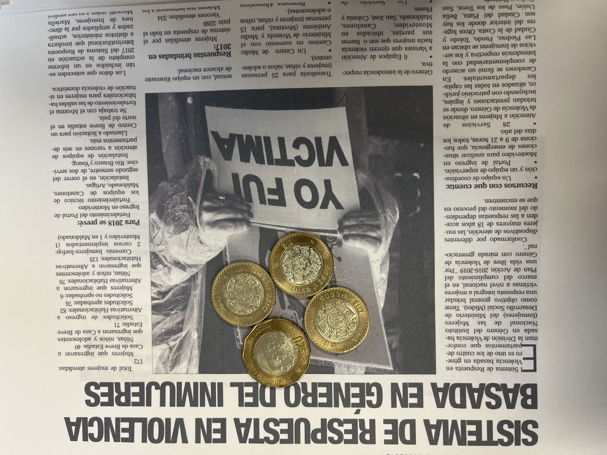 The image displays a printed document, likely a newspaper page or report, laid flat and partially obscured by four Mexican coins.
The document features a prominent black headline in Spanish: "SISTEMA DE RESPUESTA EN VIOLENCIA BASADA EN GÉNERO DEL INMUJERES" (Response System in Gender-Based Violence of INMUJERES). The accompanying text, also in Spanish, details statistics and initiatives concerning gender-based violence, specifically mentioning institutions and geographical locations within **Uruguay**, such as Montevideo, Canelones, Maldonado, Artigas, Río Branco, and Young. No locations within Puebla, Mexico, are mentioned in the visible text.
A black and white photograph is embedded within the document, partially covered by coins. It depicts the torso and arms of an unidentified individual wearing dark clothing. This person is holding a white rectangular sign horizontally across their upper body. The sign bears the capitalized black text "YO FUI VICTIMA" (I WAS A VICTIM).
Four Mexican coins are placed on the document, covering parts of the headline, text, and the photograph. There is one bimetallic $20 Mexican peso coin, identifiable by its larger size and commemorative design featuring historical figures and the years "1821 2021". Three bimetallic $10 Mexican peso coins are also present, each smaller and featuring the Aztec sun stone calendar design in the center.
The environment is limited to the printed page and the coins resting upon it. There are no discernible people other than the partial representation in the photograph, and no overt actions or interactions beyond the placement of the coins on the document.