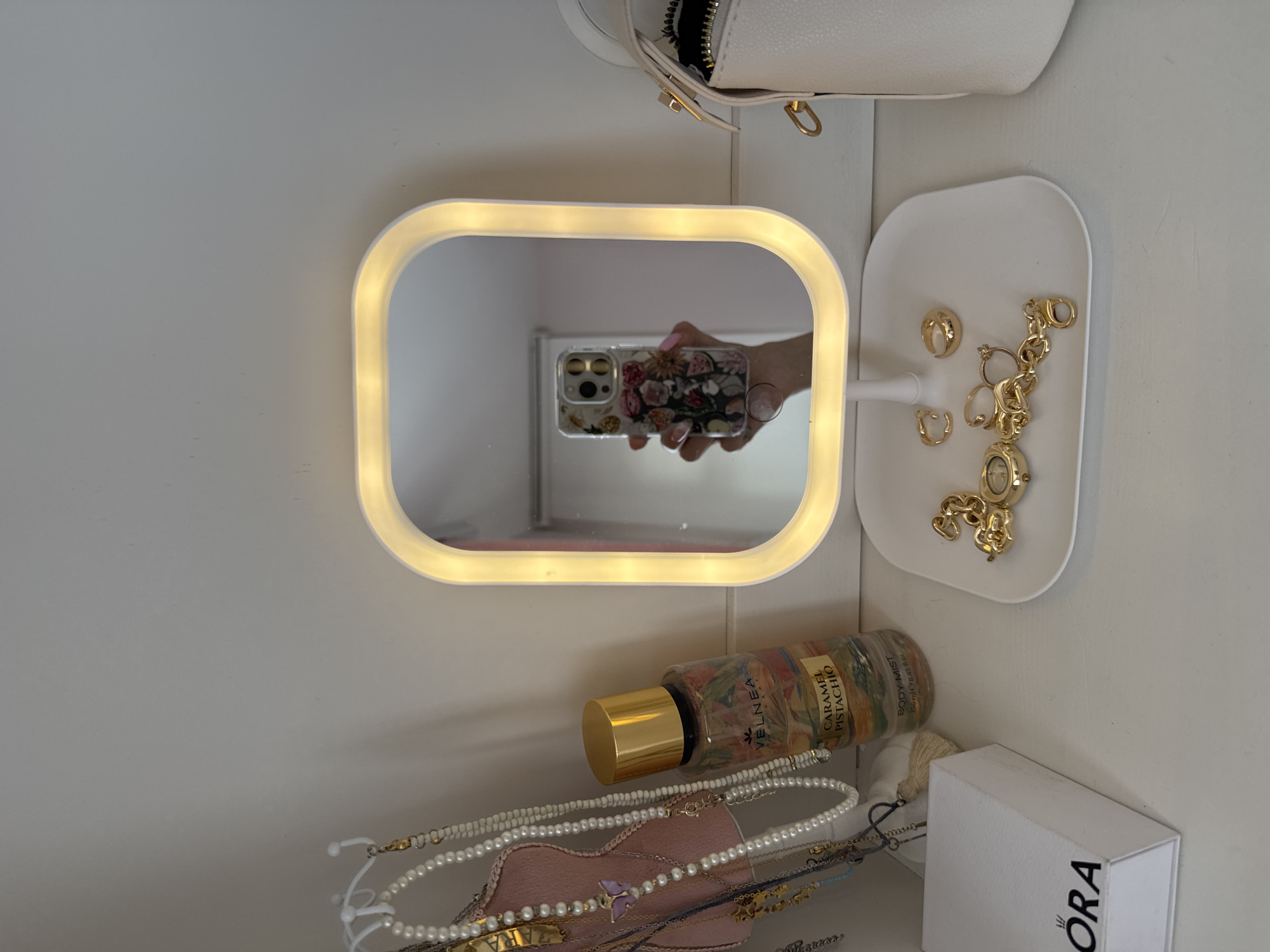 The image presents a close-up, top-down view of an organized personal space, likely a vanity or shelf, featuring a collection of accessories and beauty items. The scene is dominated by a rectangular mirror with an illuminated frame, emitting a warm, yellowish light.
Reflected in the mirror is a hand holding a smartphone, capturing the image. The smartphone has a floral-patterned case and visible camera lenses. The hand exhibits light pink nail polish.
To the right of the mirror, a white, kidney-shaped tray holds an assortment of gold-toned jewelry. This includes various earrings such as hoops, studs, and ear cuffs, along with a chain bracelet or necklace, and a small watch.
Below the mirror, a bottle of body mist, "VELNEA CARAMEL PISTACHIO BODY MIST 250ml e 8.45 fl oz," with a golden cap and a colorful label, rests on the surface. Adjacent to the body mist and partially obscured, a collection of necklaces and bracelets, including pearl strands and beaded designs, are draped over a pink fabric item, possibly a jewelry holder.
Further to the right, a white box with the brand name "PANDORA" printed in black on its side is visible. The background consists of a plain white wall or surface, providing a clean and minimalist setting for the items. The overall impression is one of personal organization and daily essentials. There are no specific visual cues to indicate the stated location context of Targovishte, Bulgaria.