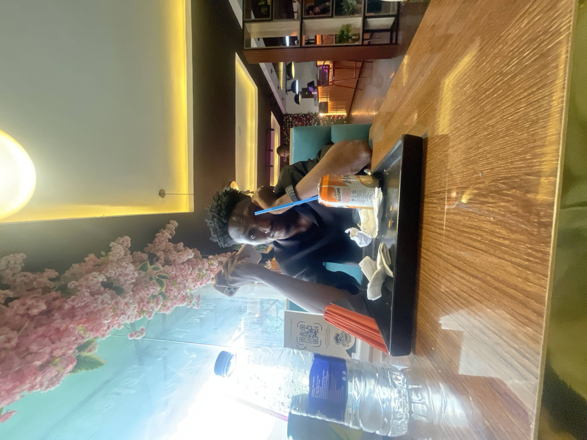 A person is seated at a table in what appears to be a restaurant or cafe. They are holding a phone and taking a photo or video. A tray on the table holds a can of Malta drink with a blue straw, a glass of juice, and some napkins. A bottle of water sits at the edge of the table. Pink artificial flowers decorate the side of a glass wall. In the background, there are other tables and chairs, and further back, shelves with decorative items. The location is in Adankolo, Nigeria.
