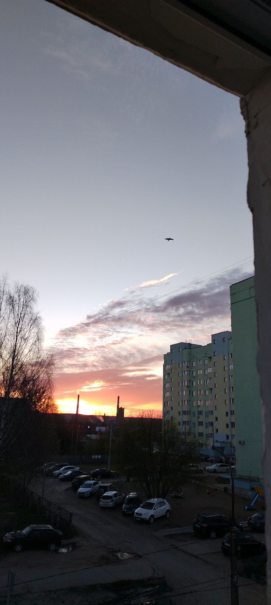 The image depicts a cityscape at sunset, framed by the edge of a building. The sky transitions from pale blue to vibrant orange at the horizon, where industrial smokestacks are silhouetted. A single bird flies high in the sky. In the middle ground, a large apartment building with numerous windows stands next to a tree. Below, a parking lot contains a collection of cars. The foreground is darker, showing the street and building details. The scene is viewed from an elevated position, likely from a window within a building. Location: Unknown, Unknown.