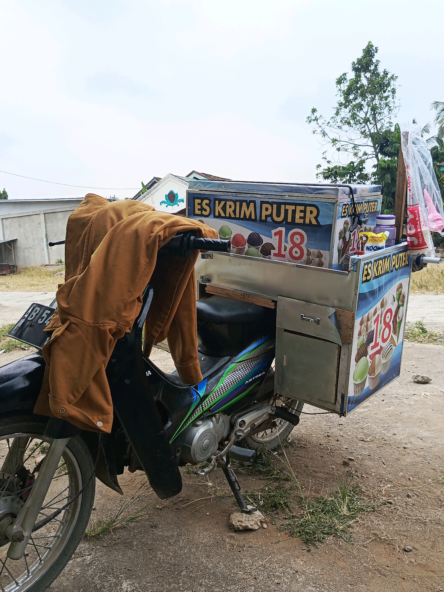 An ice cream cart is attached to a parked motorcycle. The cart is decorated with images of ice cream and the text "ES KRIM PUTER 18". A brown jacket is draped over the handlebars of the motorcycle. The scene is outdoors in what appears to be a residential area in Wonosari, Indonesia, with a building and some vegetation visible in the background. The ground is dirt and gravel. The sky is overcast.