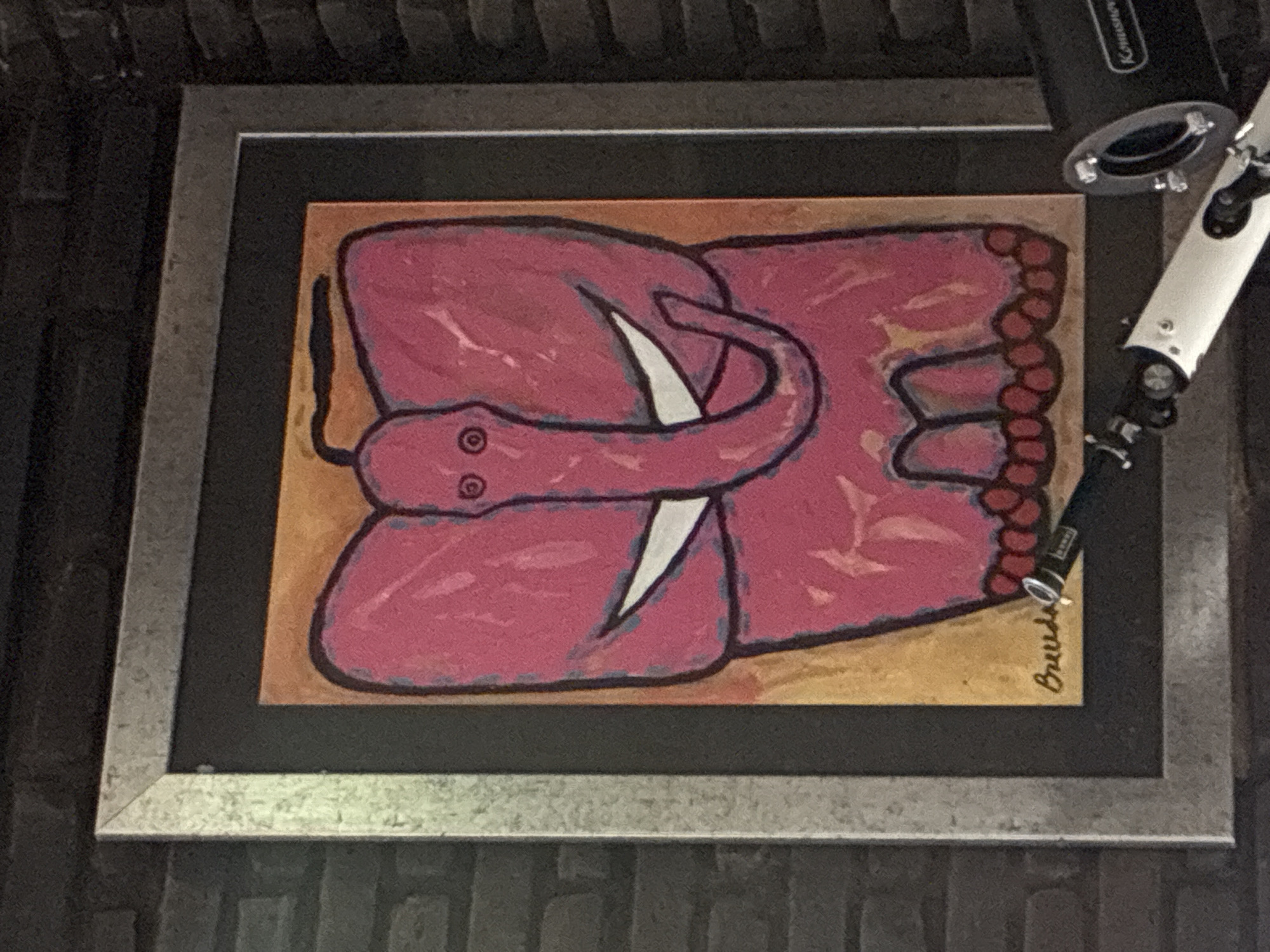 The image depicts a framed piece of artwork hanging on a textured wall. The artwork features a stylized, pink snake with two eyes and a white fang, drawn against an orange-brown background. The snake appears coiled. In the lower right corner of the artwork, there's a signature that reads "Breedlove". A black and silver frame surrounds the artwork. A black and silver object extends into the upper right corner of the frame. The image does not provide enough context to determine location in the city of Cañada de Madero, Mexico.