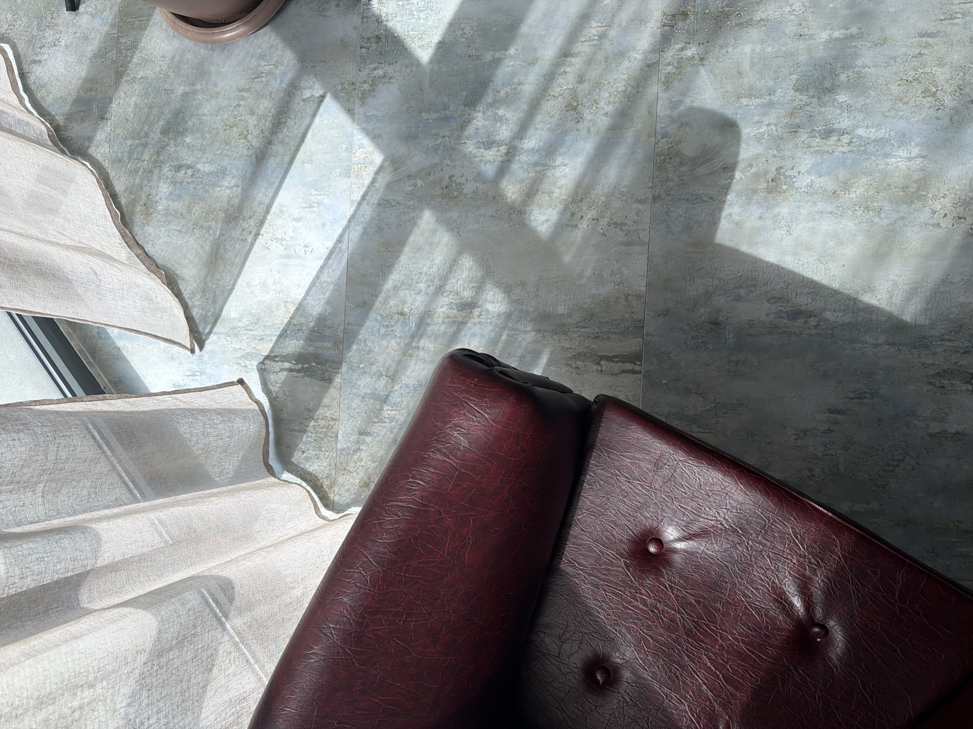 This overhead shot captures a serene indoor scene bathed in natural sunlight, set in Yerevan, Armenia. The dominant subjects include a plush, reddish-brown leather armchair, partially visible in the lower right quadrant, and sheer, light-colored curtains, cascading from the lower left corner. The armchair features distinct button tufting and a rich, textured surface.
The floor is composed of large, rectangular tiles displaying a mottled pattern in shades of light gray and subtle blue, suggesting a weathered or stone-like finish. Bright, direct sunlight streams into the room, casting strong, geometric shadows across the tiled floor, indicating either morning or late afternoon. The light also filters softly through the translucent curtains, creating gentle folds and highlighting their texture. A small, brown plant pot is partially visible in the upper left corner.
No specific activity is taking place, and no people are present, lending a quiet and contemplative atmosphere to the scene. The image focuses on the interplay of light and shadow, the textures of the furniture and flooring, and the overall peaceful ambiance of the sun-drenched interior. There is no visible text.