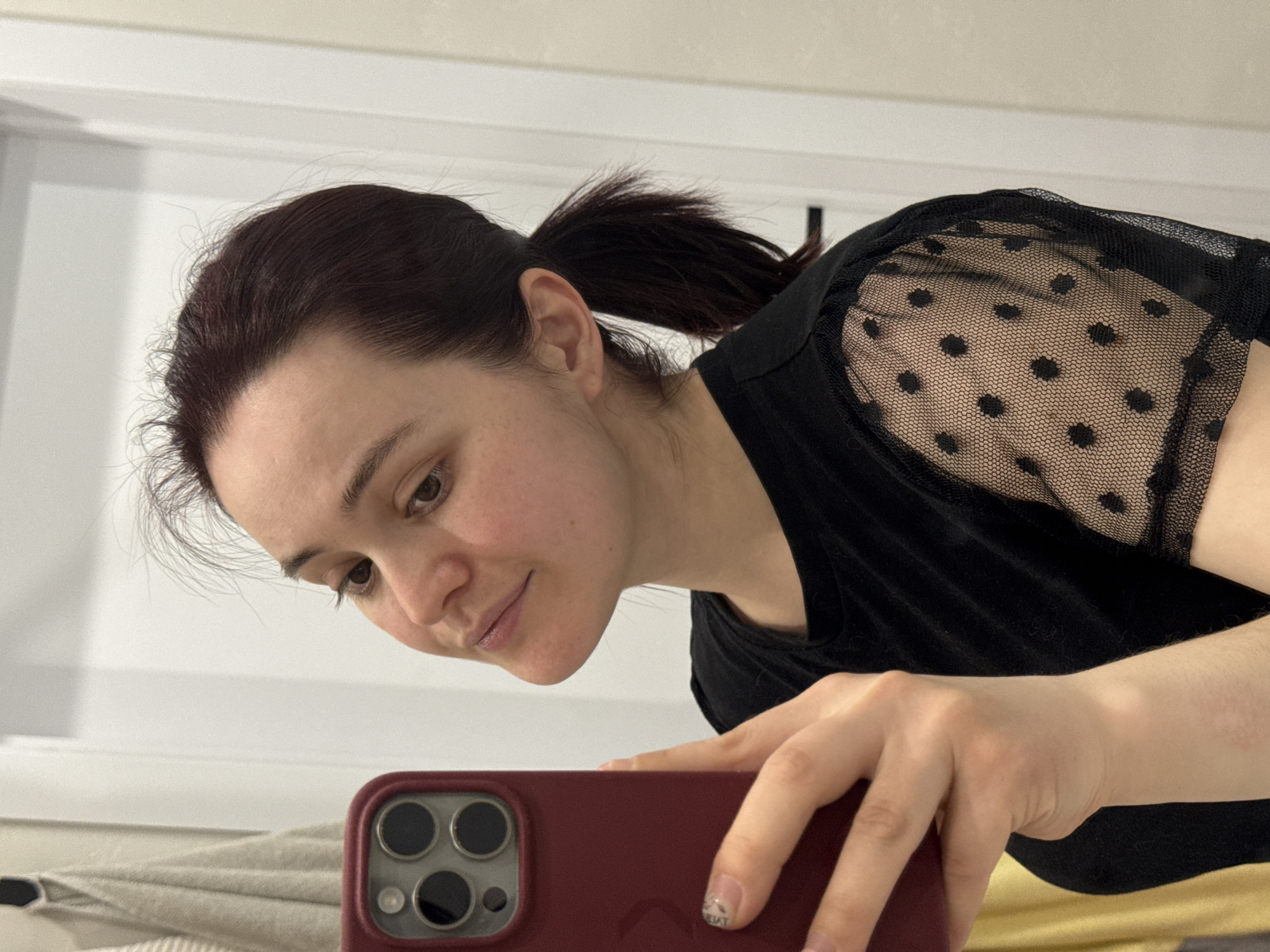 A woman with dark hair pulled into a ponytail is taking a selfie with a red smartphone. She is wearing a black top with sheer, polka-dotted sleeves. The background appears to be a room with white walls. The city of Izhevsk, Russia cannot be determined from the image.