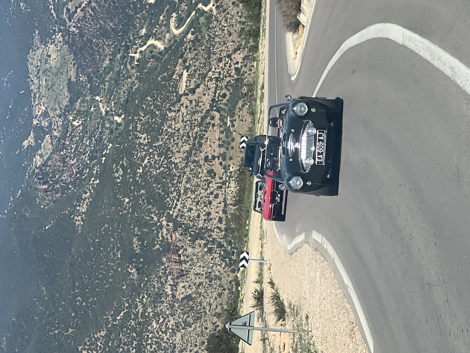 The image shows three vintage cars, one black, one dark, and one red, on a winding road next to a mountainside. The black car in the foreground has a license plate reading "EA-609-AJ". The location is identifiable as near Bin El Ouidane, Morocco, based on the mountainous terrain and the presence of classic cars on a scenic route. Two road signs are visible to the left of the cars.