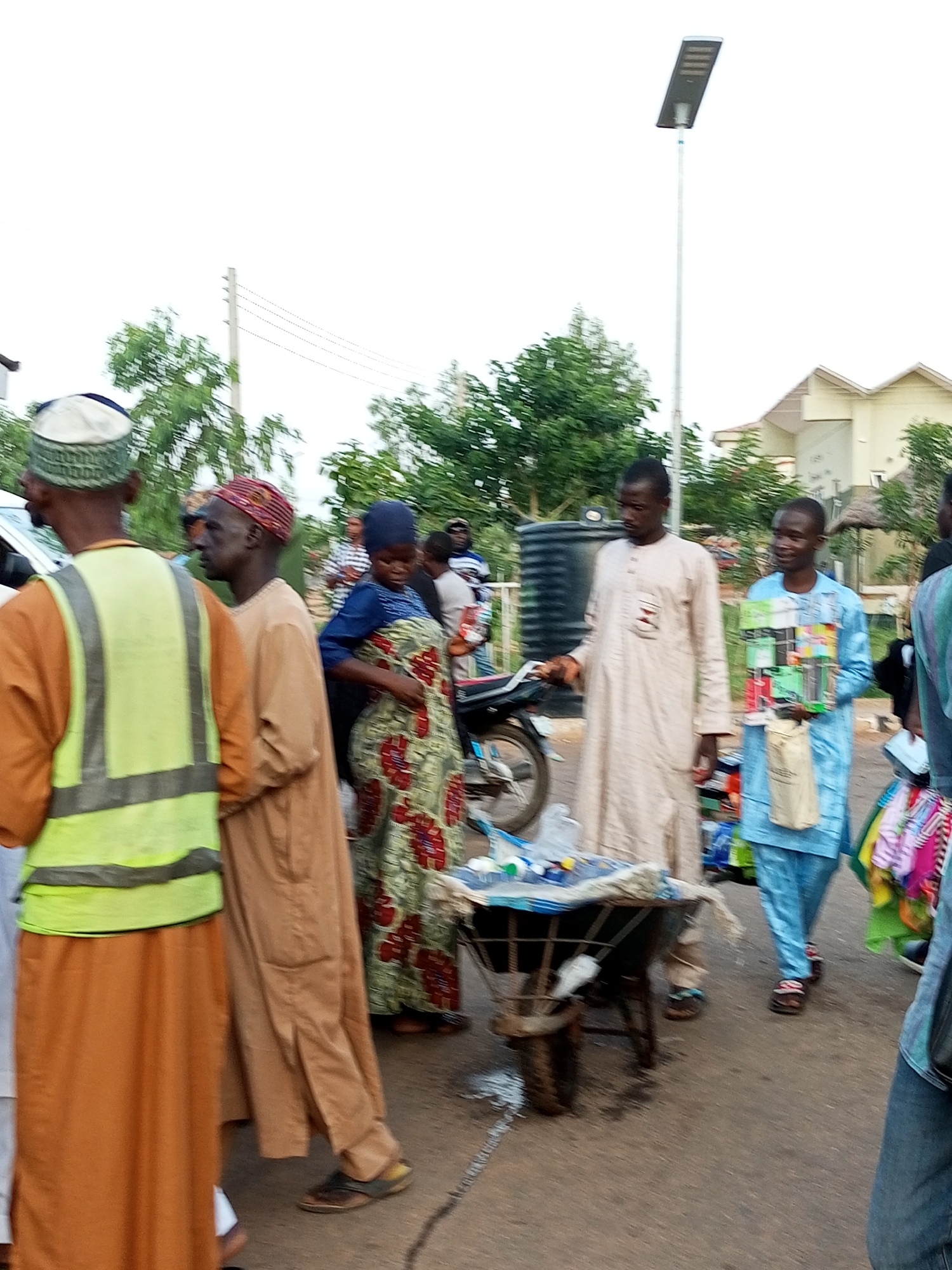 In Gombe, Nigeria, a street scene unfolds with several individuals engaged in commerce or passing by. In the foreground, a person wearing an orange robe and a high-visibility yellow vest stands facing away from the camera. To their left, another individual in a tan robe and a red and yellow patterned cap is visible. Further back, a woman in a blue head covering and a green and red patterned dress is interacting with a wheeled cart. Another man in a light-colored robe stands beside the cart, holding items. A person in a blue robe is also present, holding a collection of colorful boxes and a canvas bag. The environment appears to be a street with buildings and trees in the background, and a street lamp is prominently featured. The ground is paved.