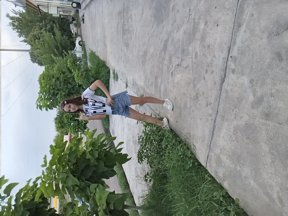 A young girl stands on a sidewalk in Angren, Uzbekistan. She is wearing a white t-shirt with black lettering, denim shorts, and white clogs. She has pink hair clips in her hair and is posing with her hand in a rock-and-roll gesture. The sidewalk is made of concrete and is cracked in places. To the left of the sidewalk is a patch of green grass and some trees. To the right of the sidewalk is a building and a street with a parked car. The sky is overcast.