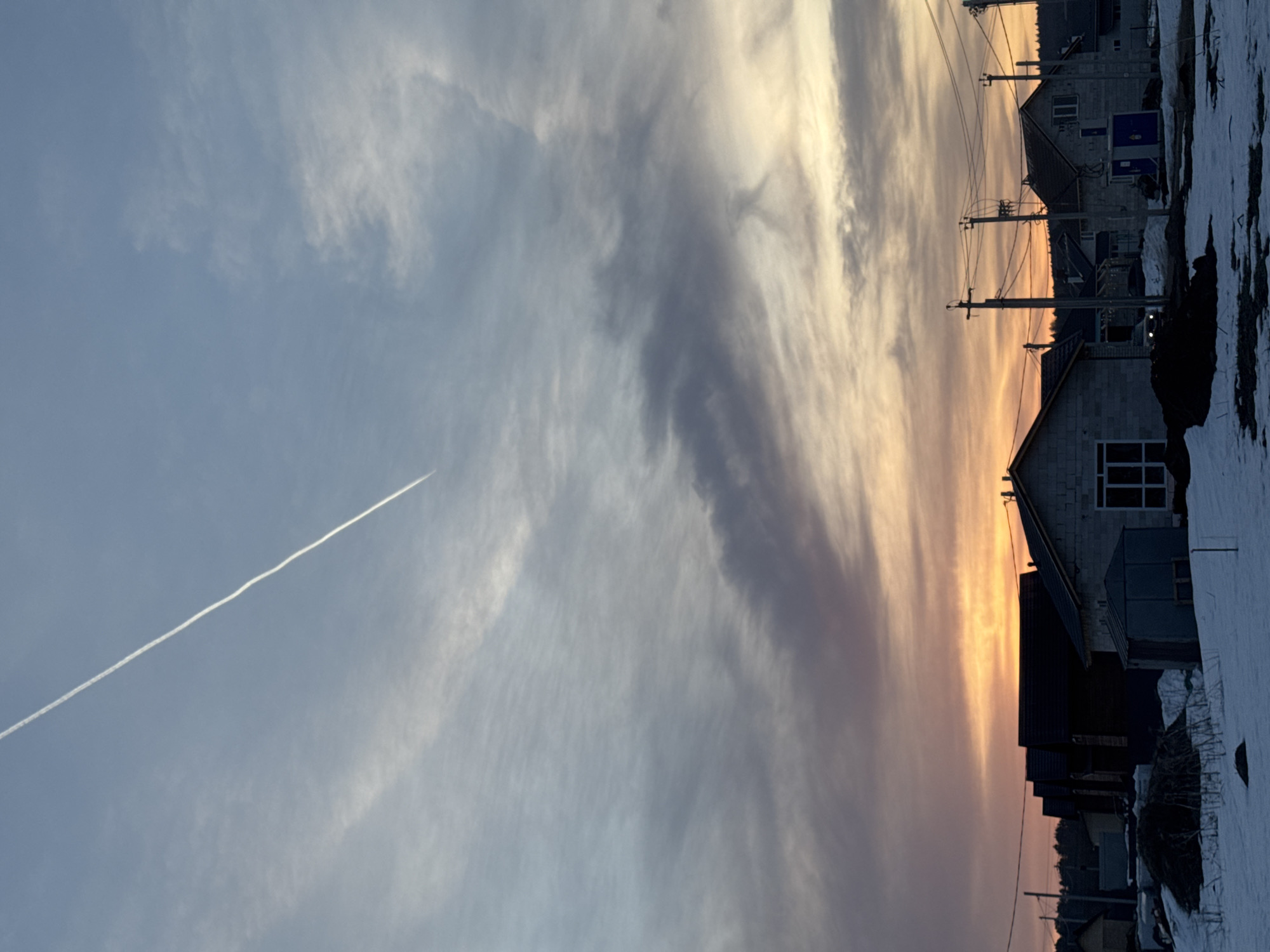 The image depicts a winter scene in Gornyi, Russia, as indicated by the visible snow. Several houses are located on the right, with a mix of gray concrete and dark roofing. Power lines run alongside the houses. The sky is a mix of light blue and orange, indicating either sunrise or sunset. Wispy clouds are visible, and a contrail from an airplane crosses the sky diagonally.