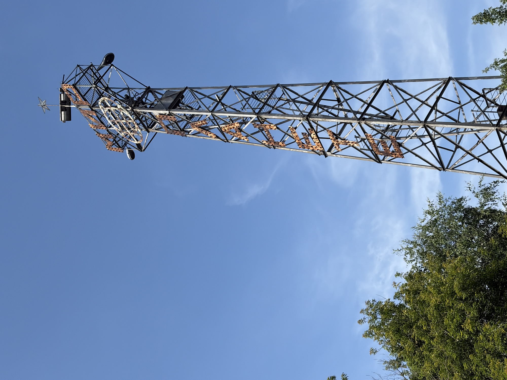 The image shows a tall, lattice steel tower against a clear blue sky. The tower is adorned with large, rusty metal letters that spell out "ЧАЙКА" (Chaika). At the top of the tower, a star-shaped ornament is visible, along with a clock face structure. The tower is located in the city of Chaikovsky, Russia, and it appears to be a prominent landmark. Trees are visible in the foreground on the left side of the image. The overall scene suggests an outdoor environment with a focus on the distinct tower.