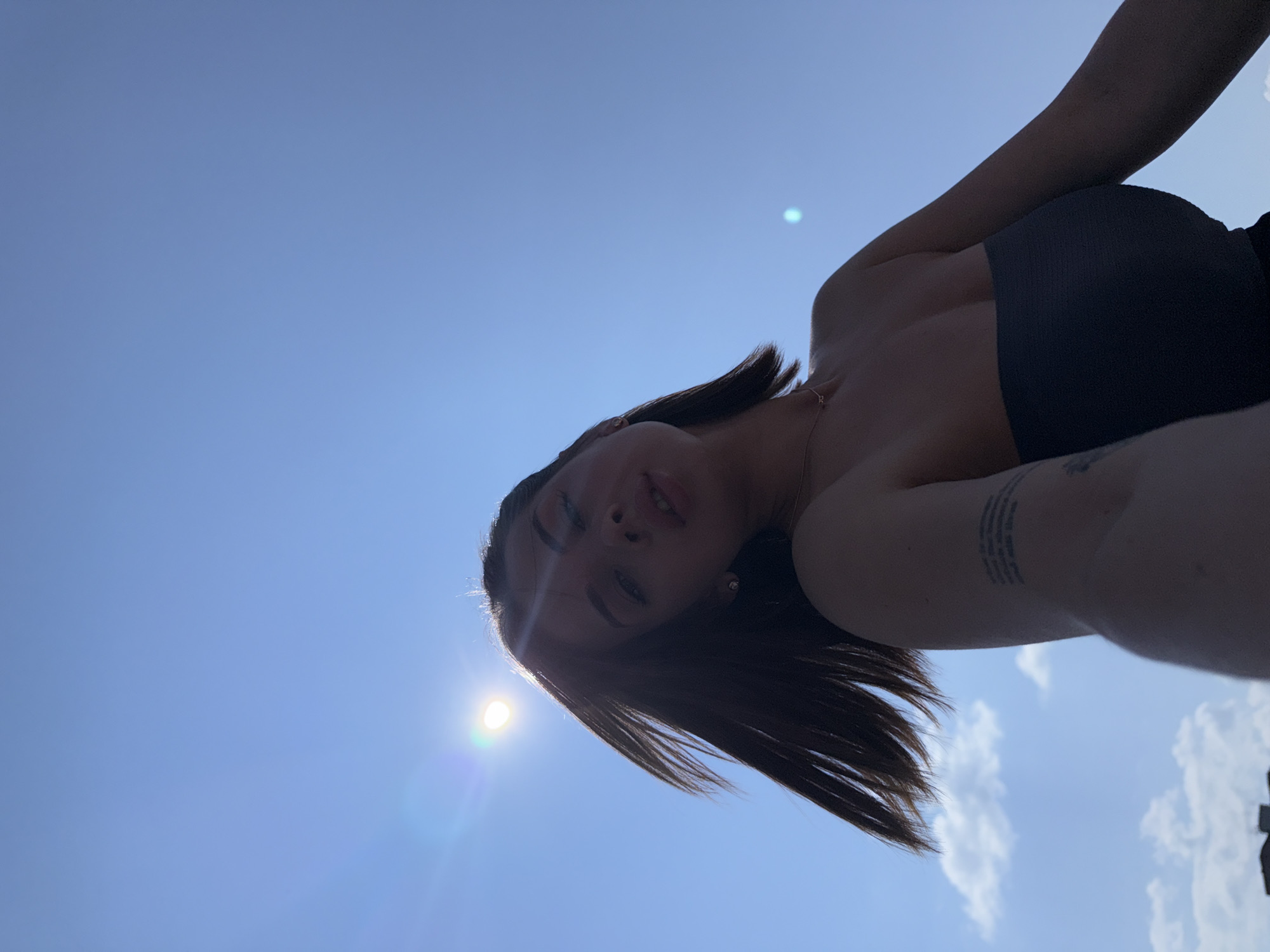 The image depicts a young woman with dark hair, wearing a strapless top, against a bright blue sky. The sun is visible, creating a lens flare. Her hair is blowing in the wind. A tattoo is visible on her left arm. The lower part of the image shows a faint outline of buildings, possibly in a city. There is no specific information to identify the location as Perm, Russia, or any other specific city.