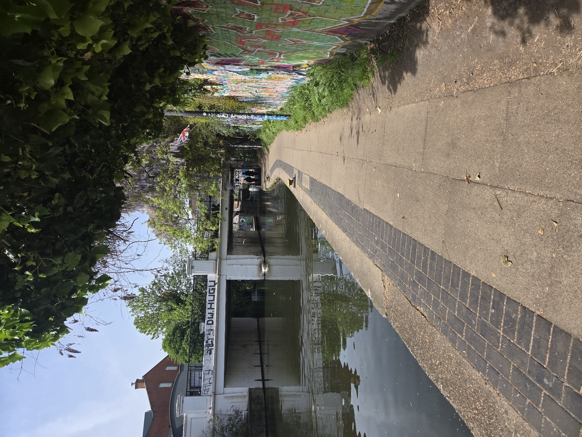 The image shows a canal in London, United Kingdom. The canal is bordered by a paved walkway on one side and a wall with graffiti on the other. The walkway is lined with trees and bushes, some of which overhang the canal. The water reflects the sky and the surrounding buildings. In the distance, a bridge spans the canal. There are a few people visible in the distance near the bridge. A Union Jack flag is visible.