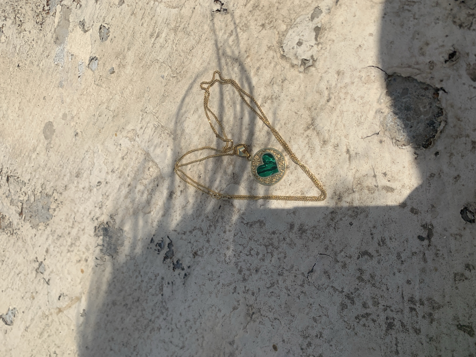 A gold necklace with a heart-shaped green stone pendant, surrounded by small clear stones, lies on a light-colored, textured surface. The surface appears to be concrete or stone with patches of lighter and darker shades, and visible cracks and imperfections. A shadow, possibly from foliage, falls across the left side of the image, creating a dappled pattern. The scene is outdoors, with harsh sunlight casting distinct shadows. There are no people or animals visible in the image. The location is specified as Khazar Raion, Azerbaijan, though this information is not visually discernible from the image itself.