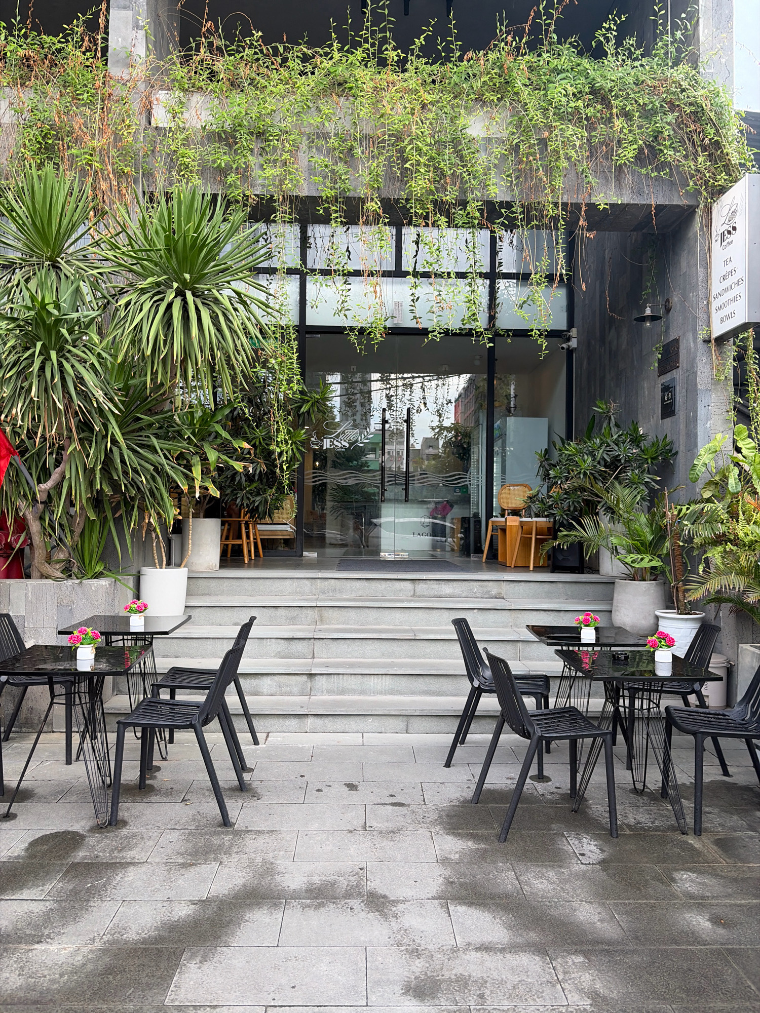 This is an outdoor photograph of a cafe in Da Nang, Vietnam. The cafe's entrance features a glass door and a few steps leading up to it. In front of the cafe, there are black metal tables and chairs arranged on a paved area. Small vases with pink flowers are placed on the tables. Green plants, including large leafy ones and hanging vines, surround the entrance and the building's facade. A sign on the right side of the entrance displays menu items like "TEA, CRÊPES, SANDWICHES, SMOOTHIES, BOWLS." The paving stones are wet in places, suggesting recent rain. The overall scene is clean and well-maintained.