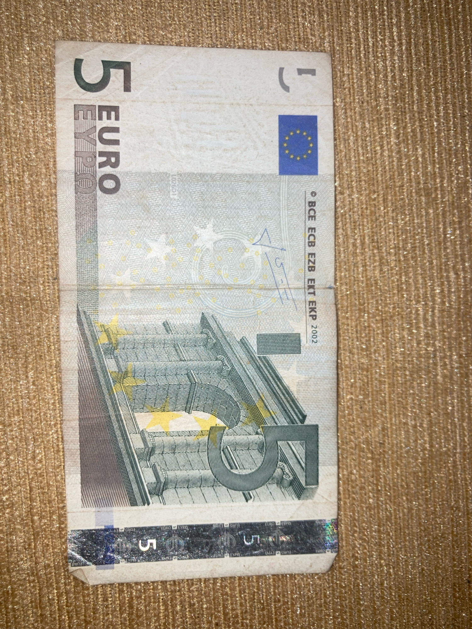 The image shows a five euro banknote placed on a textured golden surface. The banknote is folded vertically in the middle and displays the number '5' prominently in the top left corner. Below the number '5', the word 'EURO' is printed vertically. The European Union flag is visible in the upper right portion of the banknote. The reverse side of the banknote features architectural elements, resembling a bridge or a series of arches, with the number '5' integrated into the design. The banknote appears to be in good condition, with some minor creases due to the fold. The golden surface has a shimmering, fabric-like texture. There is no visible indication of the location being Daladagama, Sri Lanka; the image solely focuses on the currency and its immediate surroundings.