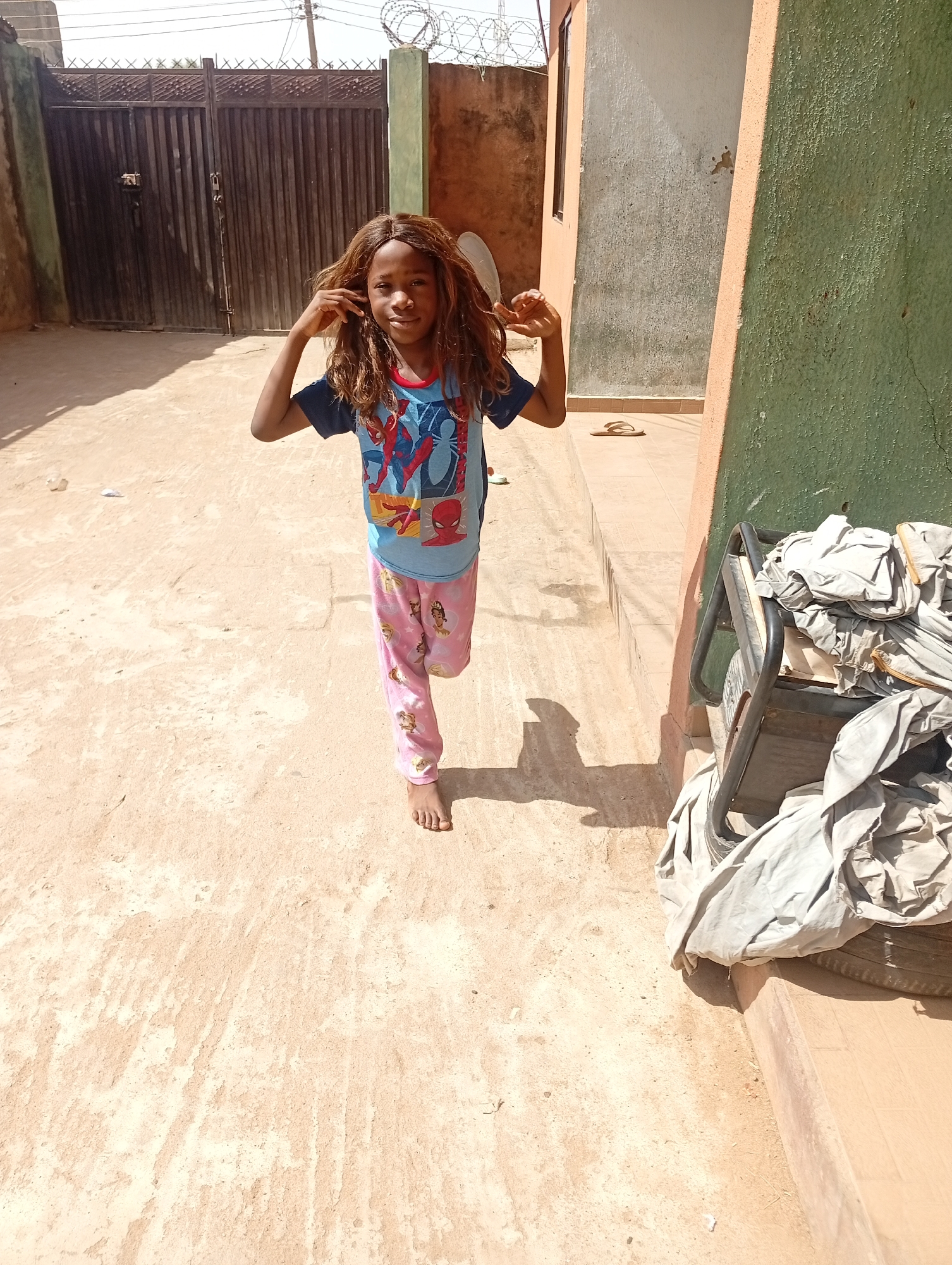 A girl stands barefoot in a sunlit, dusty courtyard in Sabon Tasha, Nigeria. She wears a blue Spider-Man T-shirt and pink patterned pajama pants. She holds her brown wig away from her face. Behind her is a dark wooden gate with barbed wire visible above it, and to her right, a wall painted green and peach. Next to the wall is a metal cart covered with folded, light-colored fabric. The courtyard surface is cracked and uneven. A pair of sandals sit on a ledge in the background.