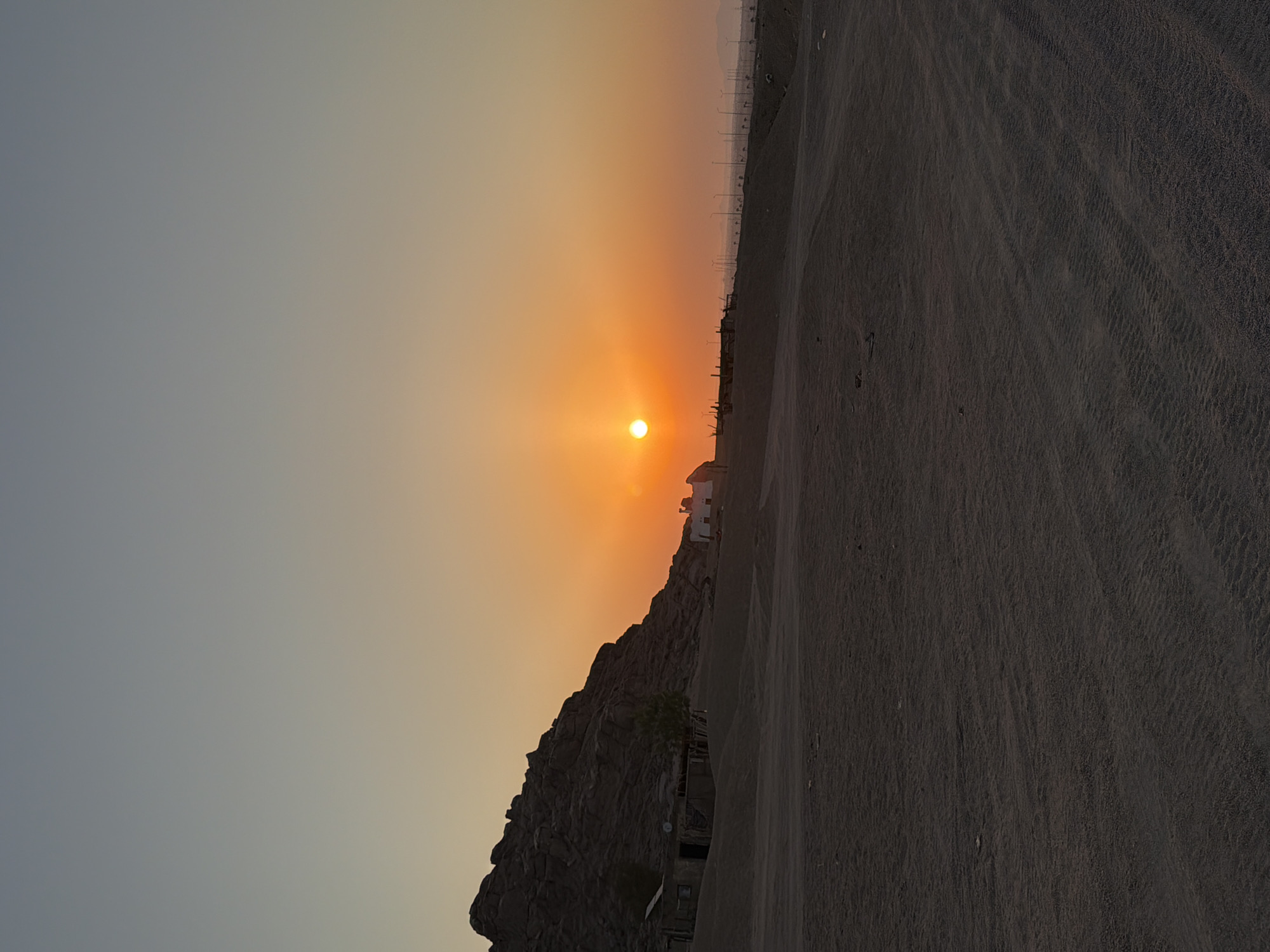 This is a landscape photograph taken during sunset in Sharm El Sheikh, Egypt. The sun, a bright orange orb, is low in the sky and casts a warm glow across the scene. The foreground is dominated by a sandy, desert-like terrain with tire tracks visible. In the middle ground, to the left, a rocky hill rises, with a few small, simple structures at its base. To the right, there are signs of development, including rows of streetlights stretching into the distance, and hints of distant mountains on the horizon. The sky transitions from a pale blue at the top to a vibrant orange near the sun.