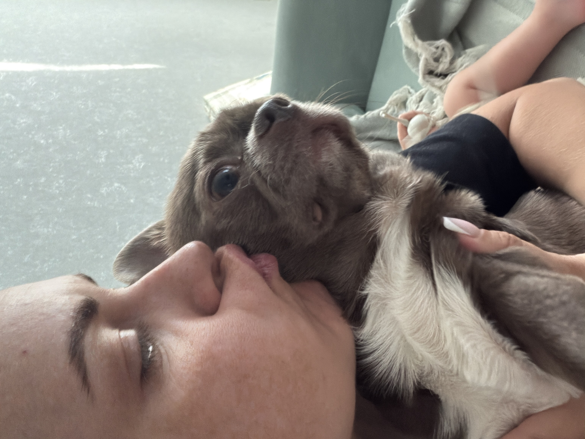 A person is lying down with their face pressed against a dog. The person's left eye is visible and closed. The dog is light brown and gray, looking up at the camera. A hand with a white-tipped fingernail is petting the dog. A blanket and a small child's legs are partially visible in the background. The background is light gray and includes a sofa. There is no information suggesting this image was taken in Votkinsk, Russia.