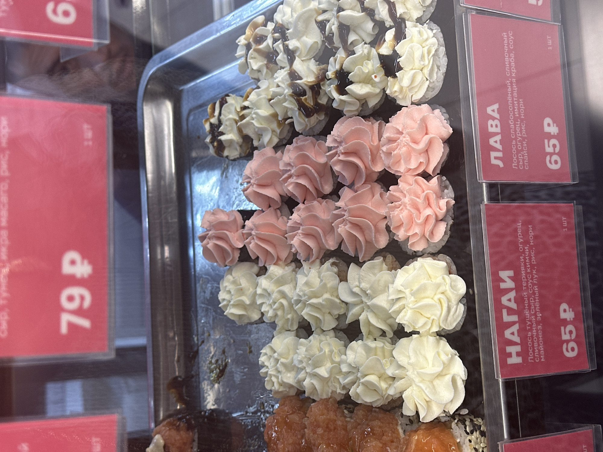 The image shows a display case filled with various types of sushi rolls at what appears to be a food establishment in Petropavlovsk-Kamchatsky, Russia. The sushi rolls are arranged in a metal tray and are topped with decorative swirls of cream cheese or frosting in white and pink. Some rolls also have a drizzle of dark sauce, possibly soy sauce or teriyaki. Price tags with Russian text and currency are visible in the foreground. One tag displays "79 ₽" and another "65 ₽". The environment appears to be indoors, likely a market or a food court, with a reflective surface in the background. There are no people directly visible in the image.