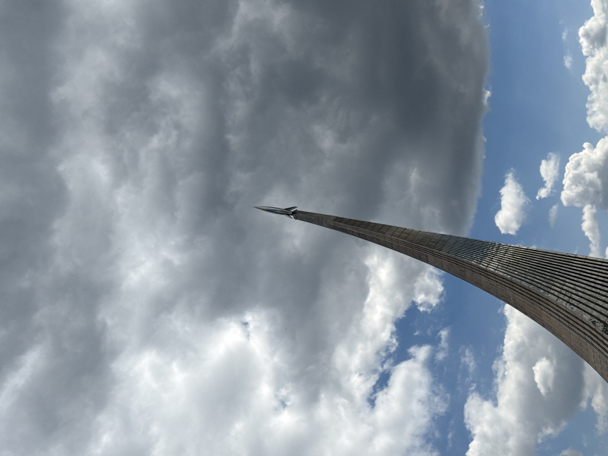 The image shows the Monument to the Conquerors of Space in Moscow, Russia. The monument features a large, curved titanium-clad obelisk topped with a model of a rocket. The sky behind the monument is filled with a mix of dark gray storm clouds and patches of blue sky with white clouds. There are no people visible in the image.