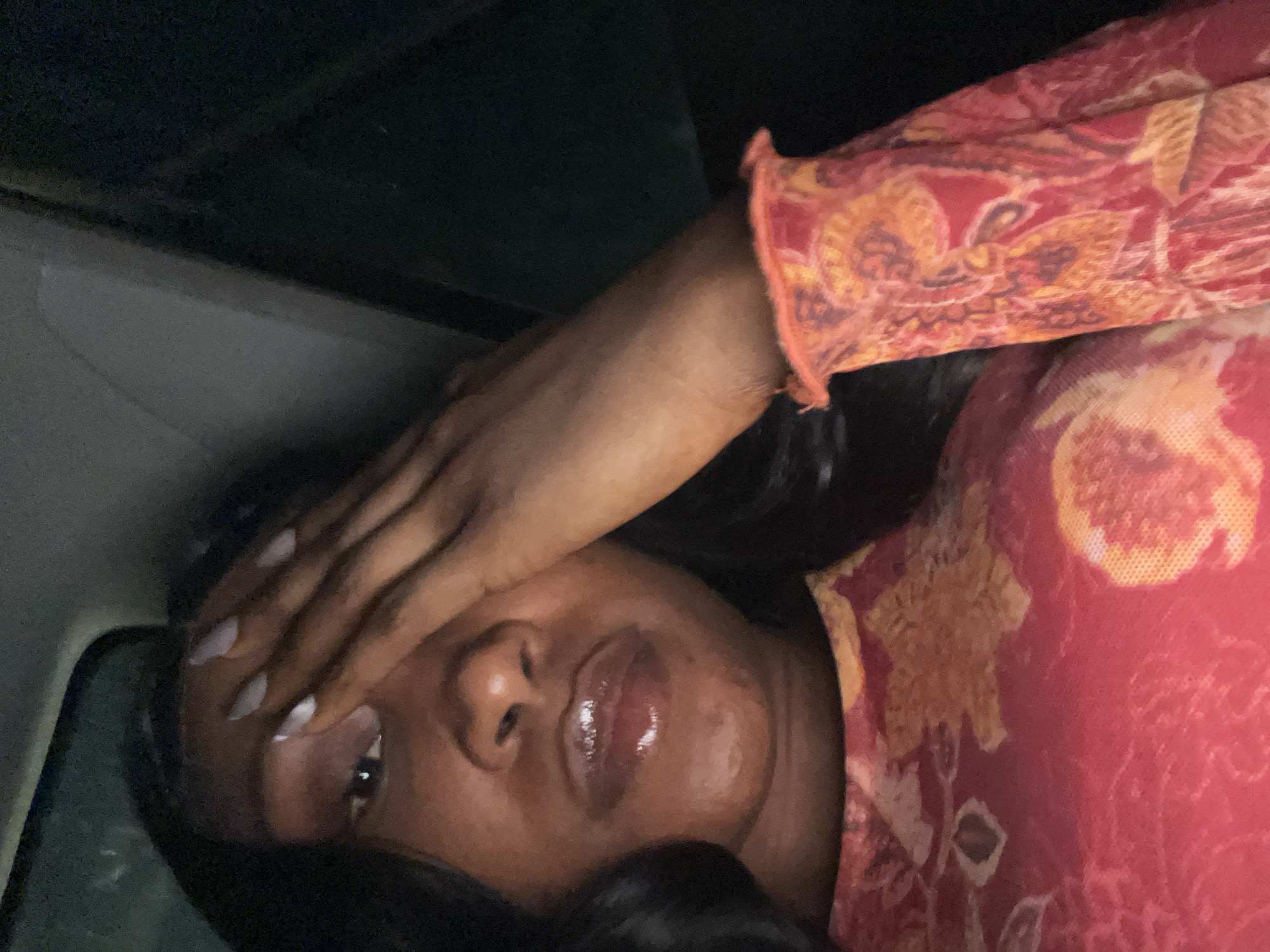 The image shows a close-up, self-taken photograph of a dark-skinned woman with long, dark hair. She is covering her face with her right hand, revealing only her left eye, nose, and mouth. Her lips are glossy. She is wearing a red top with a colorful floral print, and a ruffled cuff is visible on her right sleeve. The background is dark, suggesting a dimly lit interior, possibly a car. The image is grainy and appears to have been taken in low light conditions. The specific location in Abuja, Nigeria, cannot be determined from the image itself.
