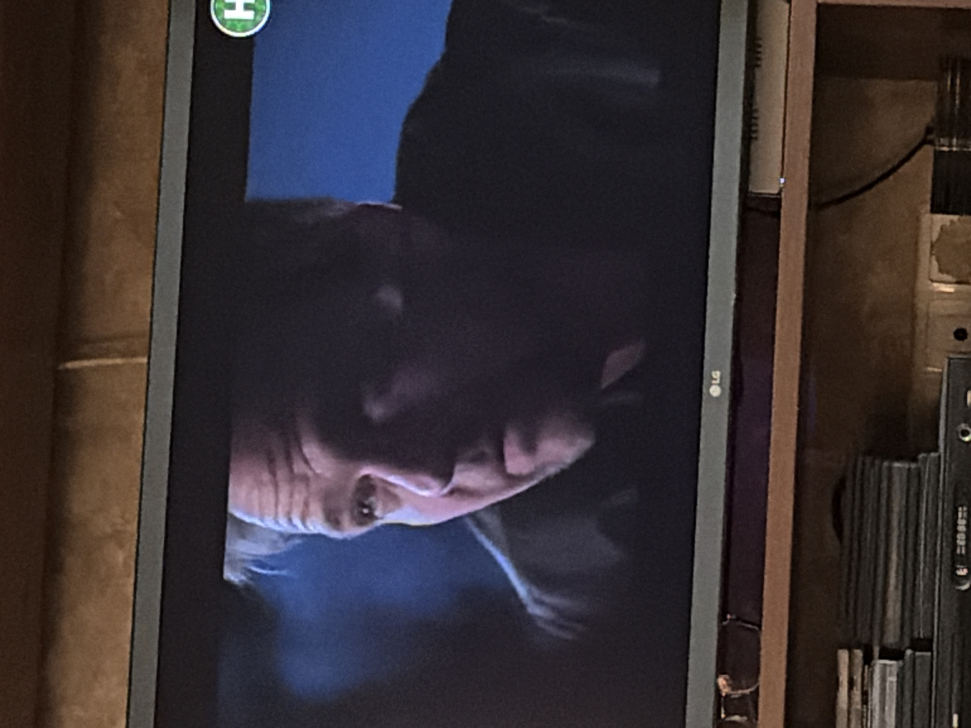 A television screen displays a close-up of a man's face, partially illuminated by blue light. The man has fair skin, greying hair, and appears to be in his late 50s or 60s. His expression is serious, with a slight frown and his mouth slightly open. He is looking off to his right. A green circle with the letter "H" inside is visible in the upper right corner of the screen. The television is placed on a wooden shelf. Below the shelf, stacks of DVDs or CDs are visible. The background behind the television appears to be a textured wall. The scene is indoors, with the lighting suggesting it is nighttime or a dimly lit room.