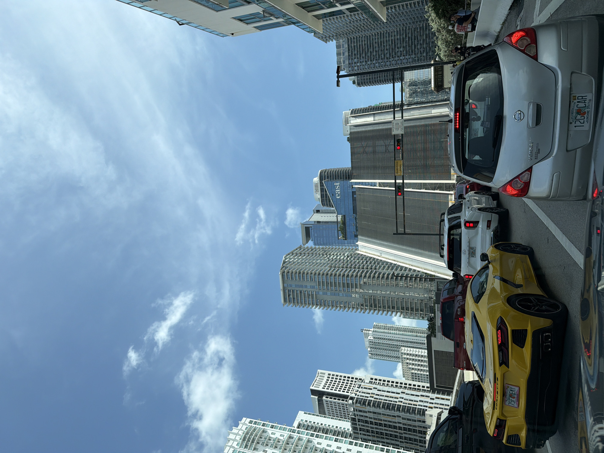 The image depicts a street scene in Miami, Florida, United States of America, characterized by a traffic jam on a sunny day. The perspective is from inside a vehicle, looking forward. Several cars are visible, including a bright yellow Chevrolet Corvette in the foreground on the left, a silver Nissan Versa in the right lane, and other vehicles further back.
The surrounding environment is urban, with numerous tall buildings that are a mix of modern architecture. These skyscrapers have distinct designs, featuring glass facades, balconies, and varying heights. Some buildings display signage, including "EAST" on one of them. A prominent feature is a large, elevated structure that appears to be a bridge or an overpass, with a ramp-like design leading upwards. Traffic lights are visible, showing red, indicating the vehicles are stopped. The sky is blue with scattered white clouds. A few people can be seen on a sidewalk in the background, near the buildings. The overall atmosphere suggests a busy urban environment during daylight hours.