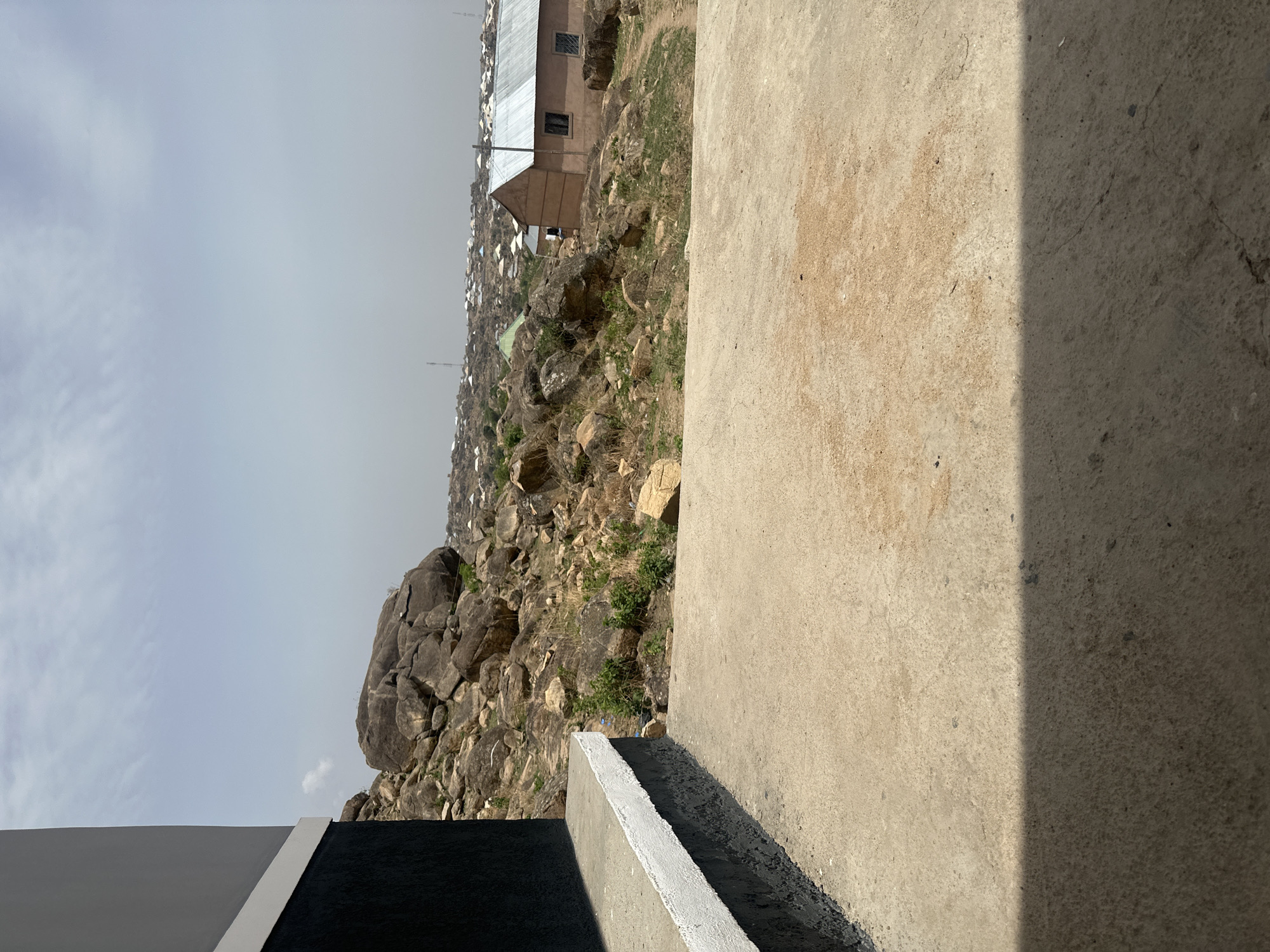 The image depicts a view of Jos, Nigeria, from an elevated concrete structure. The foreground features a concrete slab with a shadow line dividing it into two distinct lighting zones. Adjacent to this is a low concrete wall section with a painted white trim. Beyond this, a rocky landscape with sparse vegetation stretches towards the horizon. A cluster of buildings with metal roofs can be seen among the rocks, indicating residential or commercial development. The sky is overcast. No people are visible.