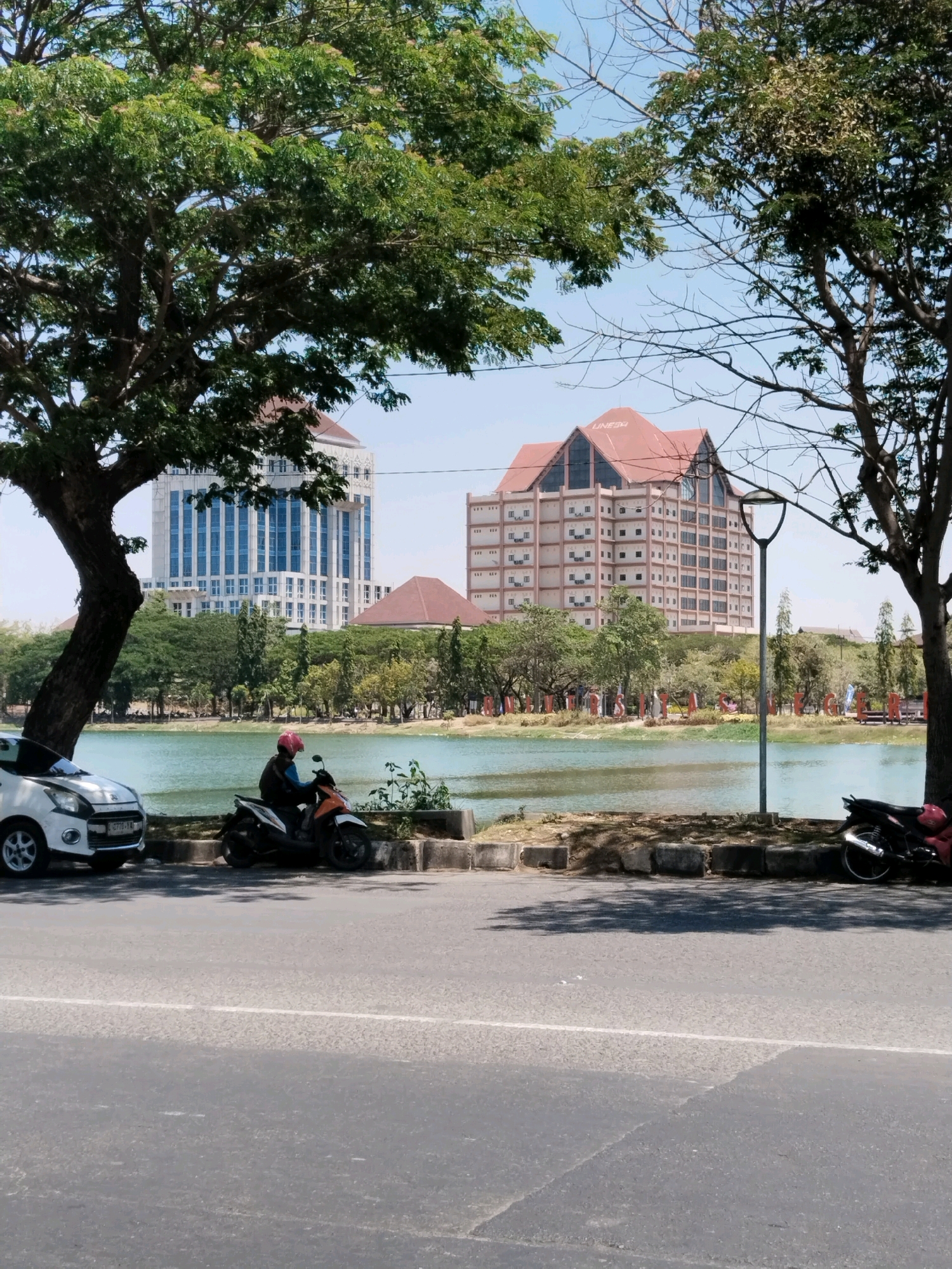 The image depicts a bright, sunny day in Surabaya, Indonesia, likely on the campus of Universitas Airlangga. In the foreground, a paved road runs horizontally across the frame, with a white line dividing the lanes. To the left, a white car is parked. Beside it, a person wearing a pink helmet and dark clothing is seated on a white and orange scooter. The scooter is parked on a raised concrete curb, which runs along the edge of a body of water.
Beyond the curb is a tranquil lake or pond, its surface a soft green. Across the lake, lush green trees line the shore, interspersed with the distinct architecture of university buildings. The most prominent structure is a large, modern building with a prominent red, A-frame roof, displaying the text "UNAIR" at its peak. To its left, another tall, multi-story building with a white facade and numerous blue-tinted windows stands. The sky above is a clear, pale blue. The overall scene conveys a sense of an academic campus environment on a pleasant day.