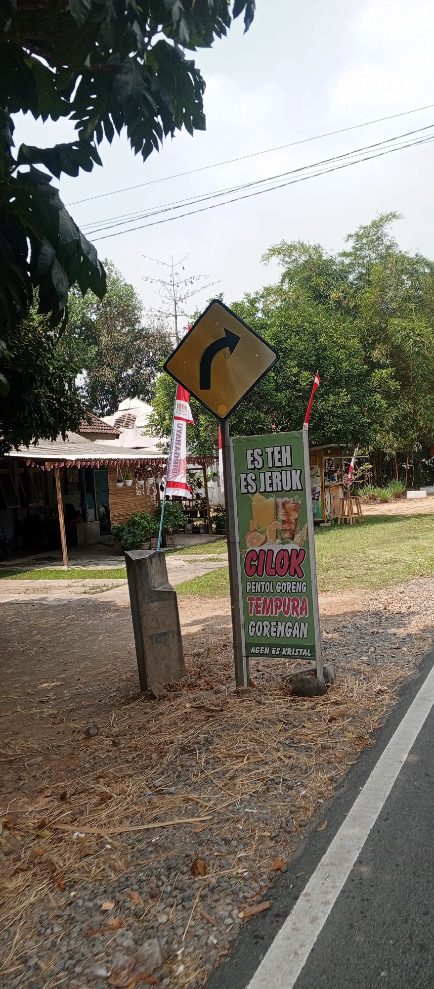 The image depicts an outdoor scene in Maospati, Indonesia, featuring a roadside stall advertising food and drinks. In the foreground, a paved road with a white line marking its edge is visible, bordering dry grass and gravel. A concrete block stands to the left. A prominent green sign with white and red lettering advertises "ES TEH ES JERUK," followed by "CILOK PENTOL GORENG TEMPURA GORENGAN AGEN ES KRISTAL," indicating beverages and fried snacks are sold. Behind the stall, a building with a red-tiled roof is partially visible, with flags and greenery surrounding it. A yellow diamond-shaped road sign with a black arrow curving to the right is mounted on a pole, suggesting a turn ahead. In the background, lush bamboo trees and other vegetation form a dense natural screen, under a bright, cloudy sky. The overall impression is of a typical Indonesian roadside eatery in a semi-rural setting.