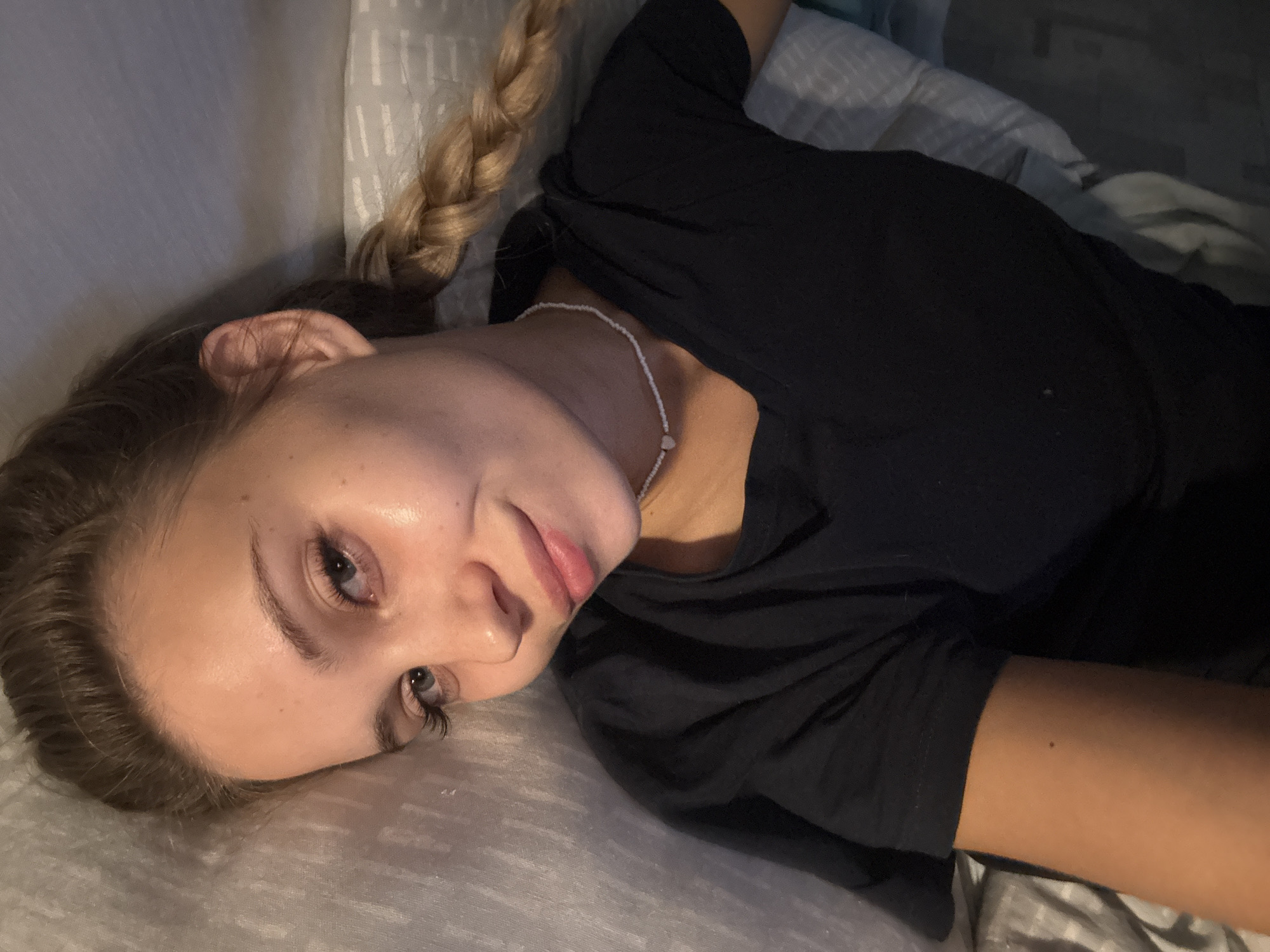 A young woman with blonde hair styled in a braid lies on a grey pillow or cushion. She wears a black shirt and a white beaded necklace. She is looking at the camera with a slight smile. The background appears to be bedding with a textured pattern. There is no information to associate this image with the city of Chaikovsky, Russia.