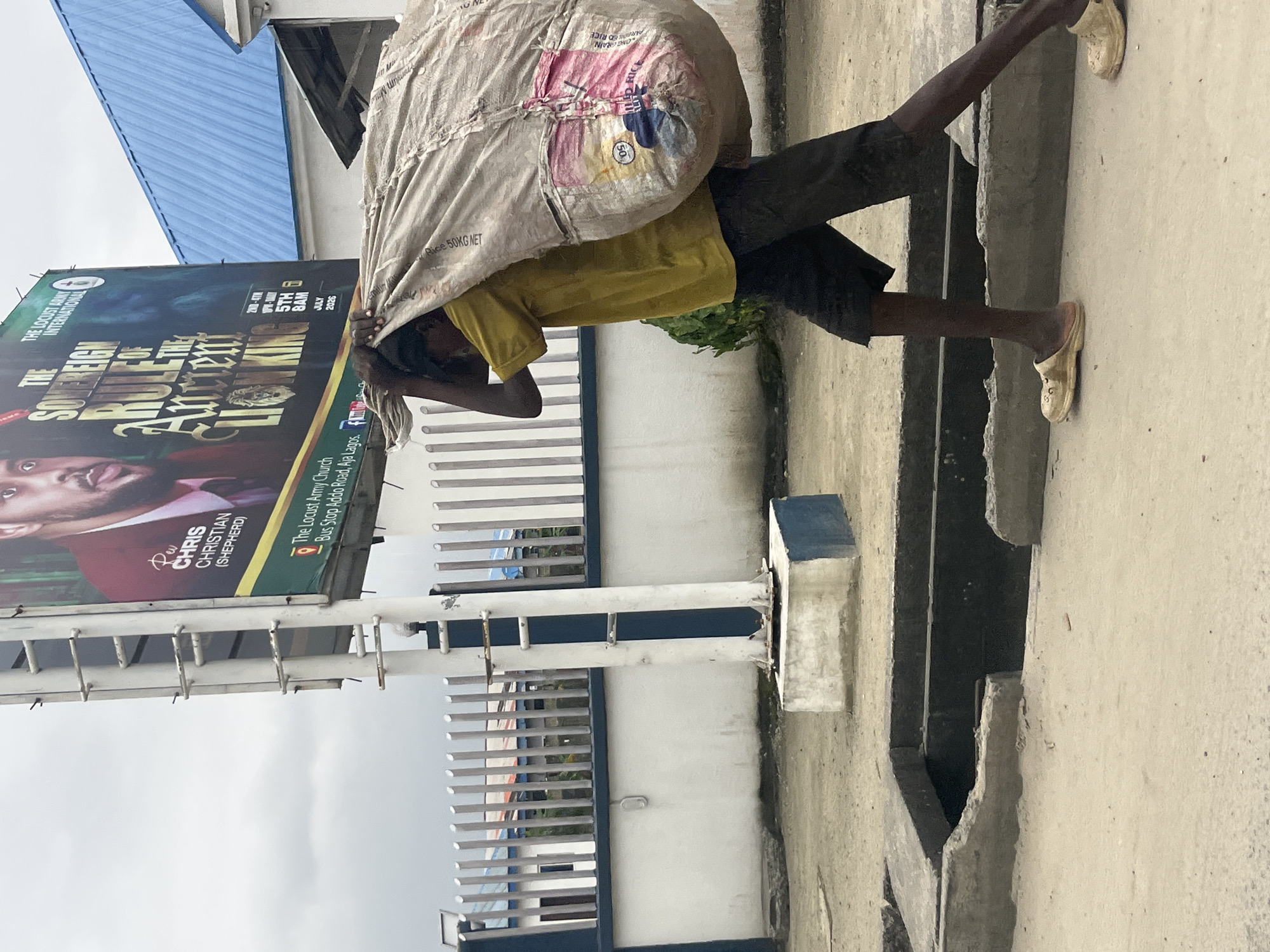 A person, likely male based on physique and attire, walks in a street in Ado, Nigeria, carrying a large, lumpy sack over their right shoulder. The sack appears to be made of a rough, woven material and has visible pink and white markings, one of which displays the number "50" and the text "KG NET". The person wears a yellow t-shirt, dark shorts, and light-colored slip-on shoes. Their left hand is raised, gripping the top of the sack for support. The person is walking on a sandy, paved surface next to a concrete drainage ditch that runs parallel to the street. In the background, a tall billboard stands prominently, advertising "THE SOVEREIGN RULE OF THE ANCIENT LION KING" with text below indicating "Rev. CHRIS CHRISTIAN (SHEPHERD)" and "The Locust Army Church, Bus Stop Addo Road, Aja Lagos." Adjacent to the billboard is a building with a blue corrugated metal roof and white walls, bordered by a metal fence. The sky is overcast.