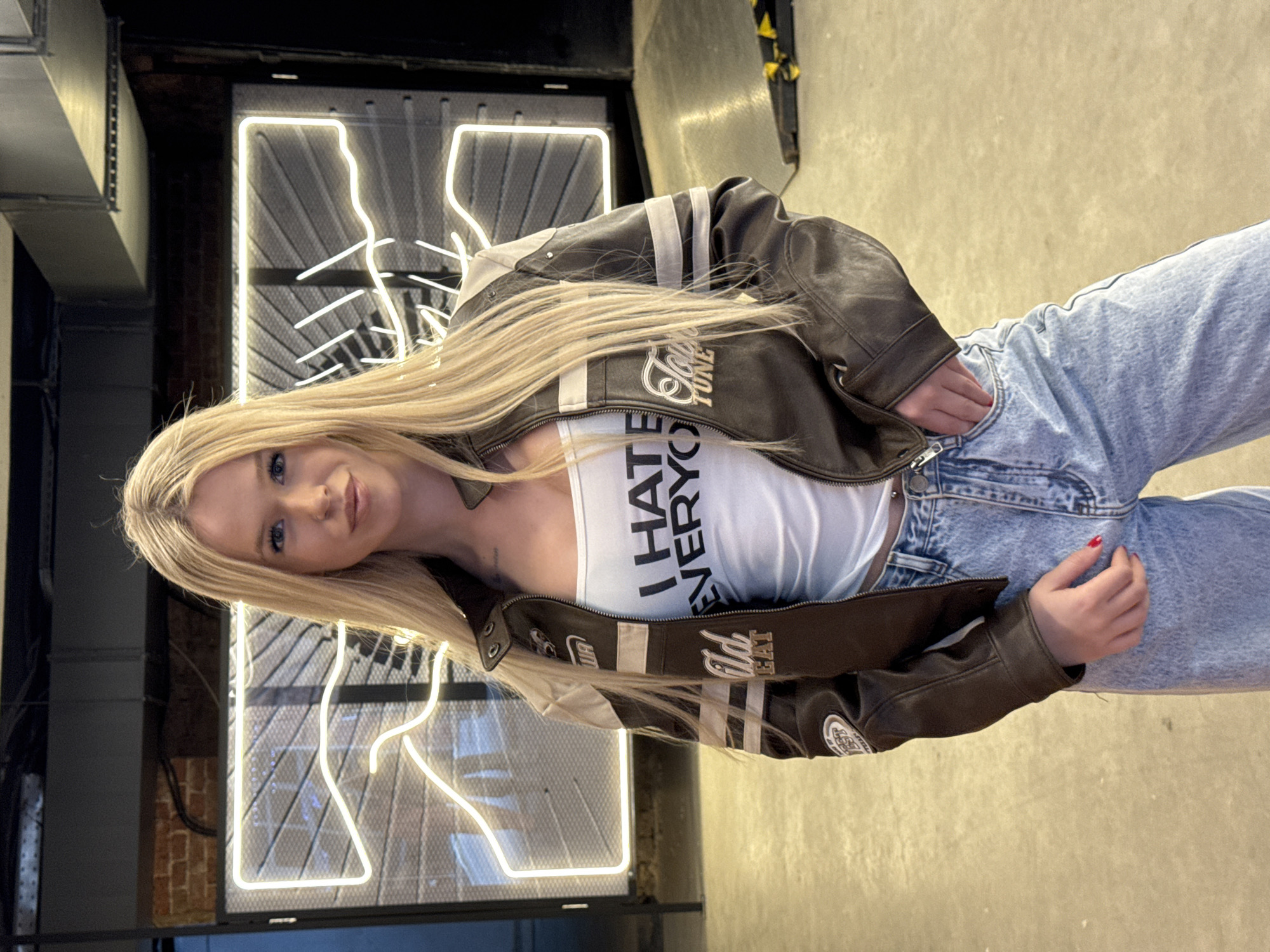 A young woman with long blonde hair is posing for a photo in a dimly lit industrial-style space. She is wearing a brown leather jacket with white stripes and patches, a white tank top with the words "I HATE EVERYONE" printed in black, and light-wash distressed jeans. Her left hand is casually tucked into her jeans pocket, and her right hand is resting on her hip.
Behind her, a large, illuminated neon sign in the shape of the letter "K" glows brightly against a dark, textured wall. Industrial elements, such as exposed brick and metal ductwork, are visible in the background, suggesting the location is a warehouse or a similar urban setting. The overall mood is casual and confident, with the woman looking directly at the camera with a slight smile. The lighting from the neon sign casts a soft glow on the scene. There is no indication of the time of day or weather conditions. The provided location context is Saint Petersburg, Russia.