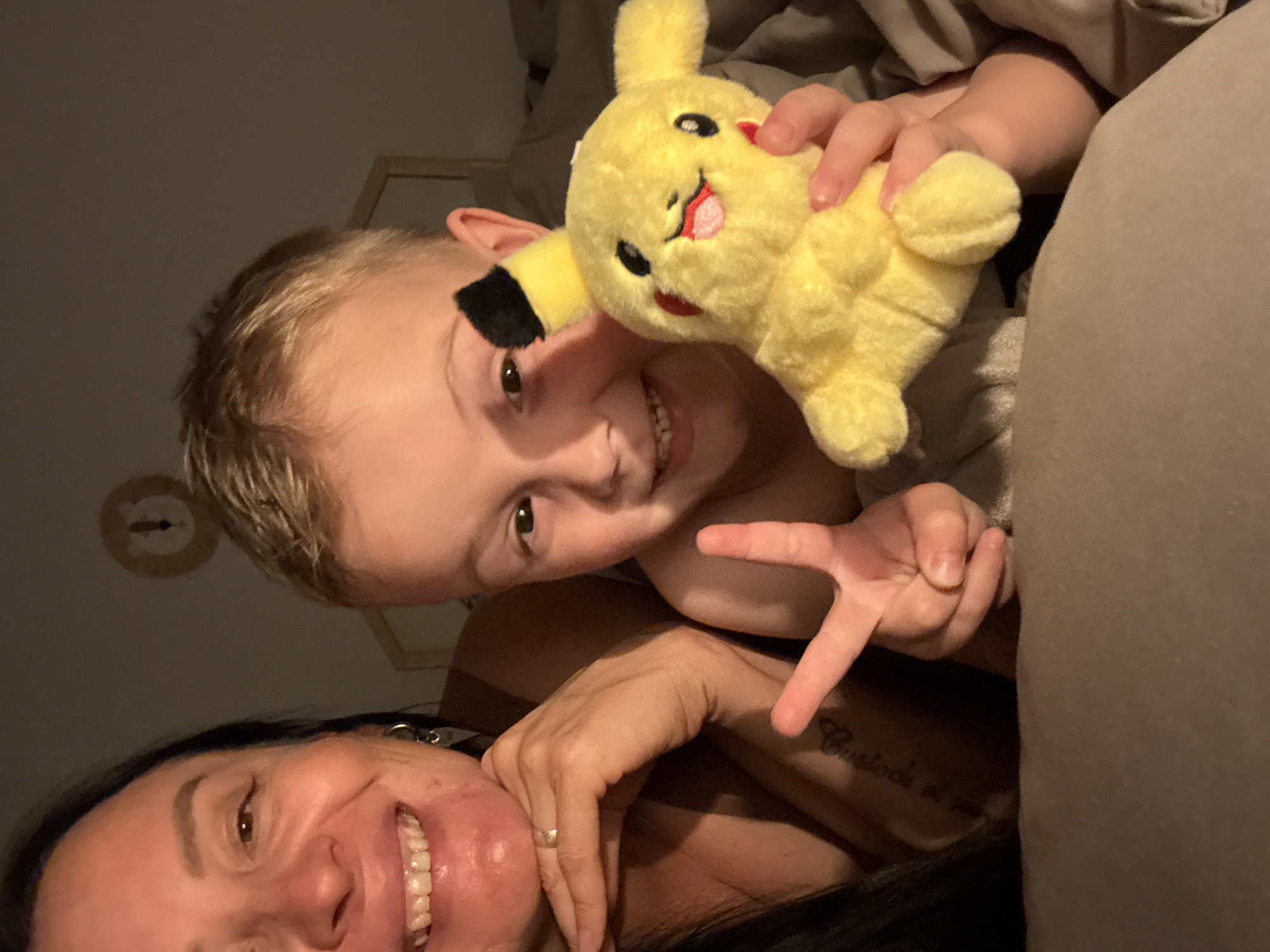 A selfie captures a woman and a young boy lying down, likely in an indoor, domestic setting. The boy holds a Pikachu plush toy. He makes a V sign with his fingers and smiles. The woman is smiling with her teeth showing. The image is angled slightly, and the lighting is subdued. A tattoo is visible on the woman's arm. The location is unspecified, and there is no indication of Chaikovsky, Russia, in the image.