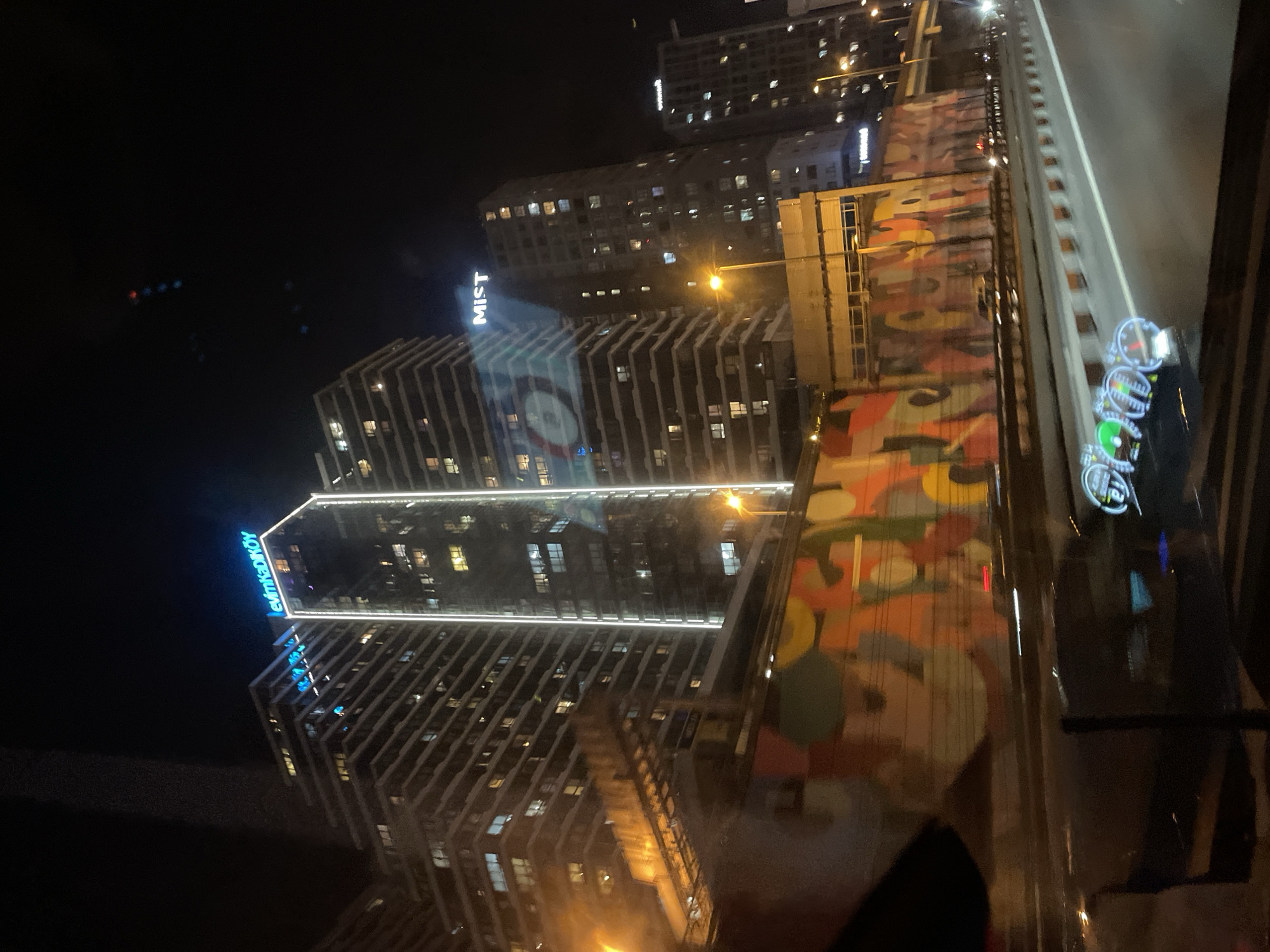 This image captures a nighttime view of the Kadıköy district in Istanbul, Turkey, as seen from a moving vehicle. Dominating the scene are several modern high-rise buildings, illuminated by exterior lights and the warm glow of interior windows. One prominent building displays a neon blue sign that reads "evinkadiköy," identifying the location. Another building features a white sign that says "MIST." In the foreground, a highway or major road is visible, with car headlights and taillights creating streaks of light due to the vehicle's movement. A vibrant, abstract mural adorns a wall or structure alongside the road. The overall atmosphere is urban and dynamic, characteristic of a city at night.