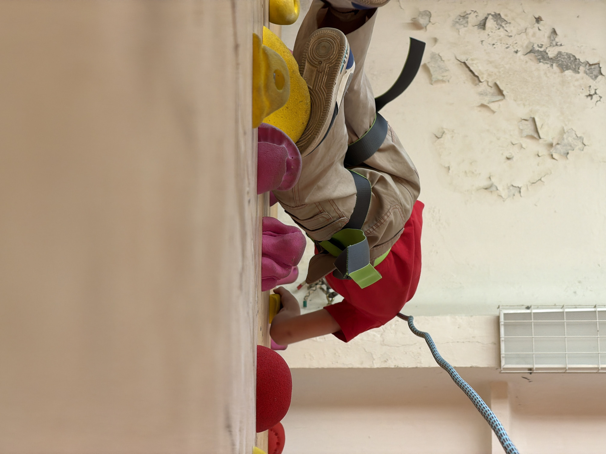 A child wearing a red shirt and tan pants is climbing an indoor rock climbing wall in Izhevsk, Russia. The child is wearing a climbing harness and climbing shoes. The climbing wall has various colored holds. The background shows a peeling wall and a rope extending from the child's harness.