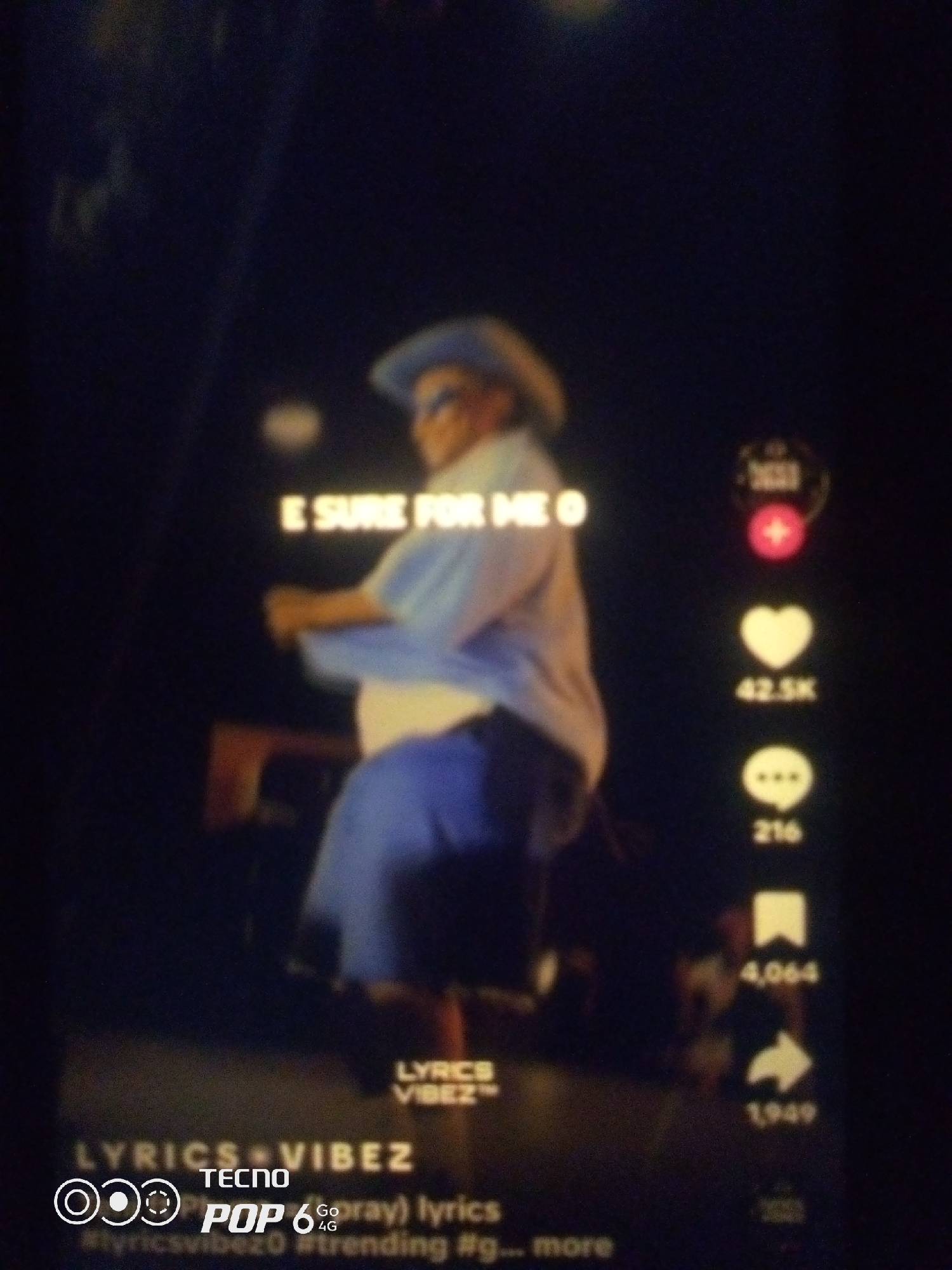 The image is a still frame from a video, likely a social media post, displaying a person wearing a hat and a light blue shirt with a white undershirt, dancing. The person's lower body is clad in blue shorts. The background is dark, with some illuminated elements suggesting a stage or performance setting. On the right side of the frame, there are typical social media interaction icons: a heart with a count of "42.5K", a speech bubble with a count of "216", a bookmark with a count of "4,064", and a share icon with a count of "1,949". At the bottom of the image, text overlays indicate "LYRICS VIBEZ", "TECNO", and "POP 6 Go 4G". The text also includes hashtags such as "#lyricsvibezo", "#trending", and "#g...more". There is no visible indication of the city of Howayi, Nigeria, within the image itself. The image quality is blurry, suggesting it was taken from a screen or in low light conditions.